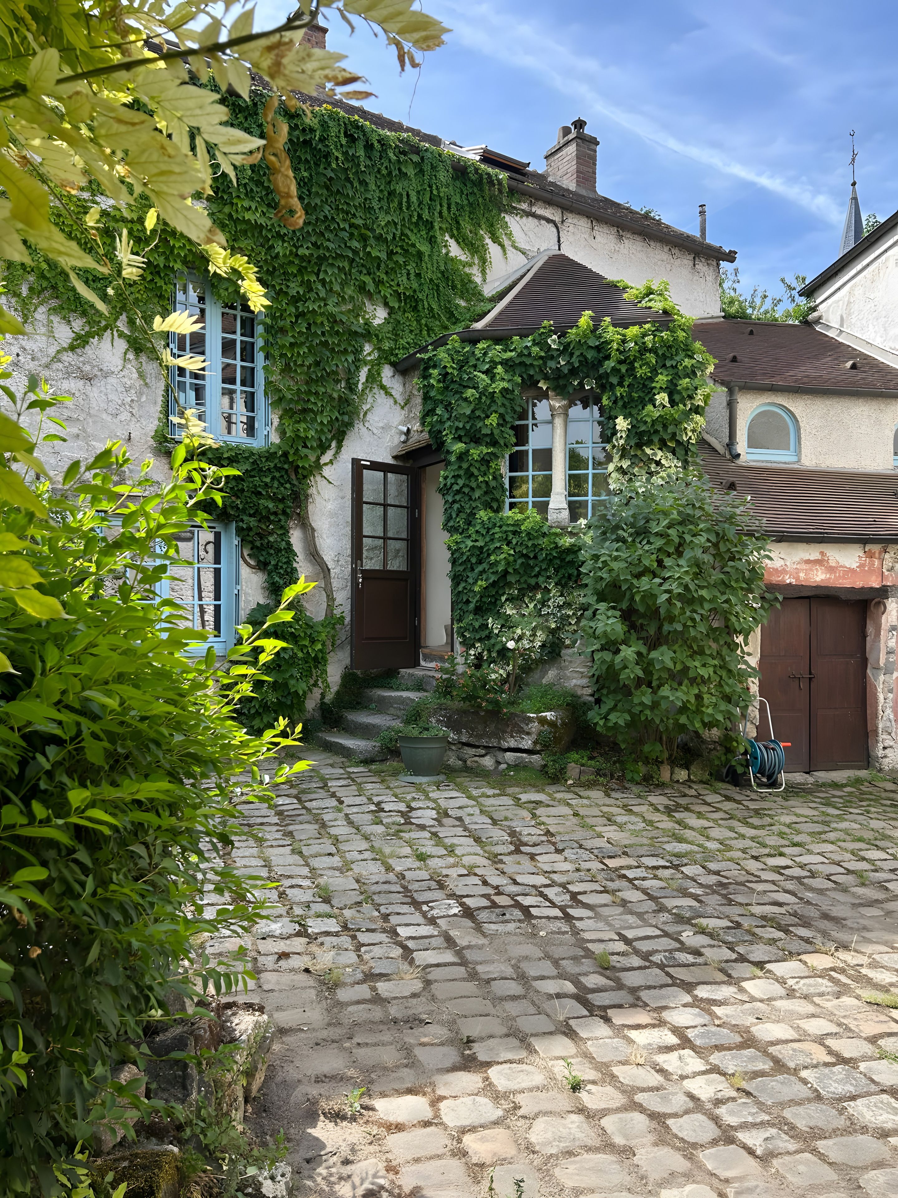 A cobblestone courtyard with a building covered in ivy, a wooden door, and a small garden area.
