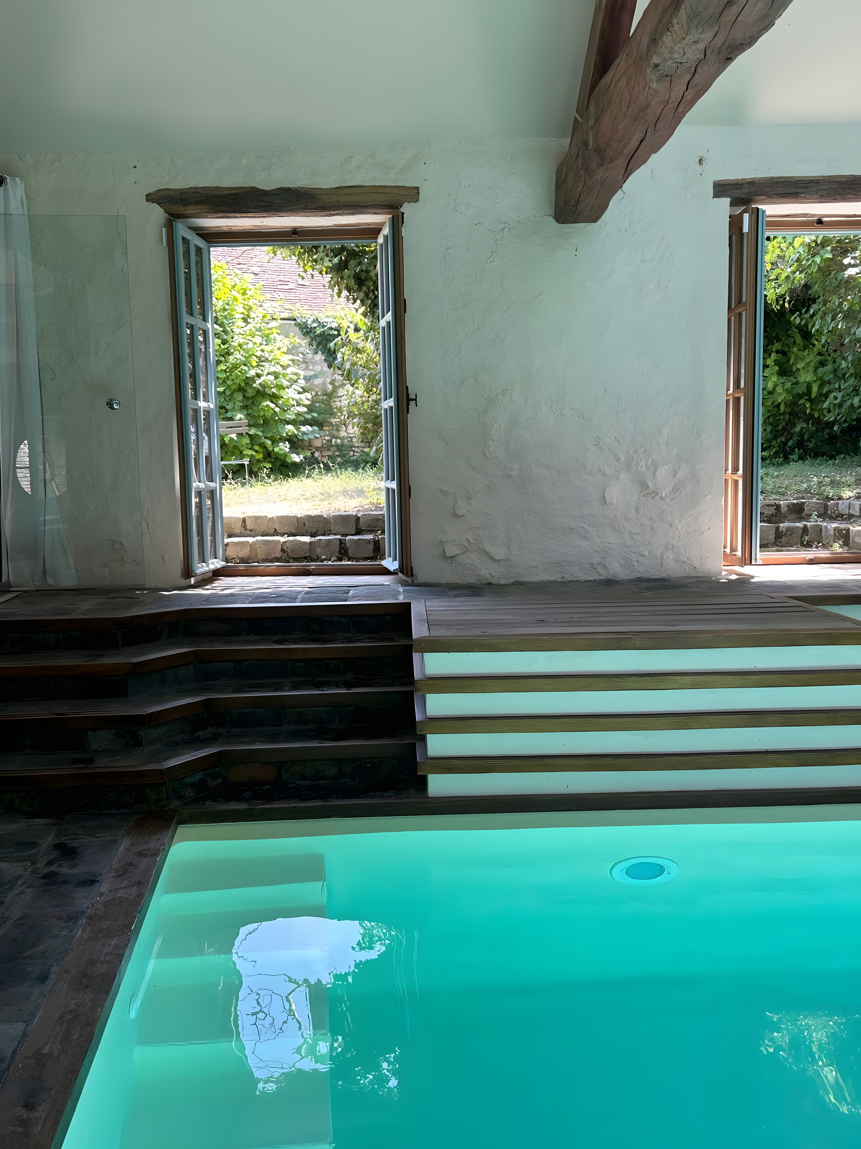 Una piscina con escalones que la bordean, rodeada por un muro blanco con vigas de madera.