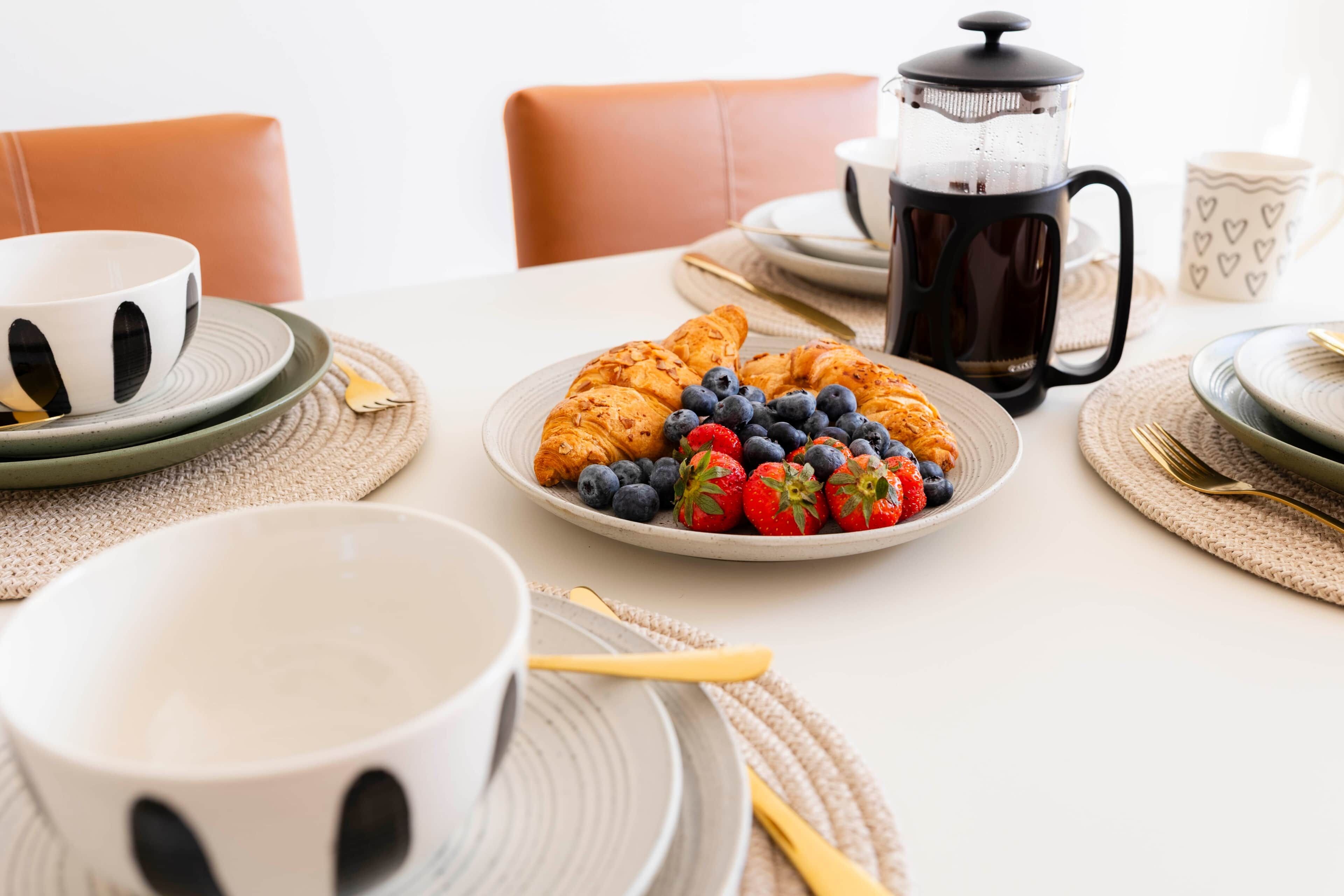 A breakfast setting with croissants, strawberries, and blueberries on a plate, a coffee pot, and a cup.