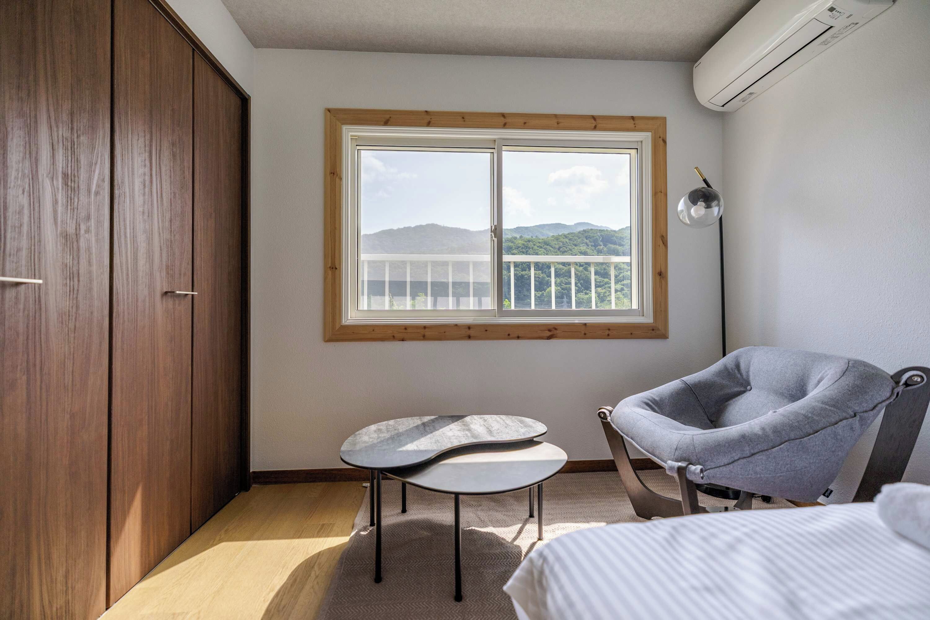 A bedroom with a large window, a wardrobe, a chair, a small table, and a bed.