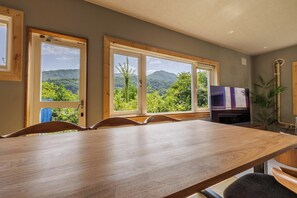 A dining area with a long wooden table and chairs, large windows with a mountain view, and a television on the wall.