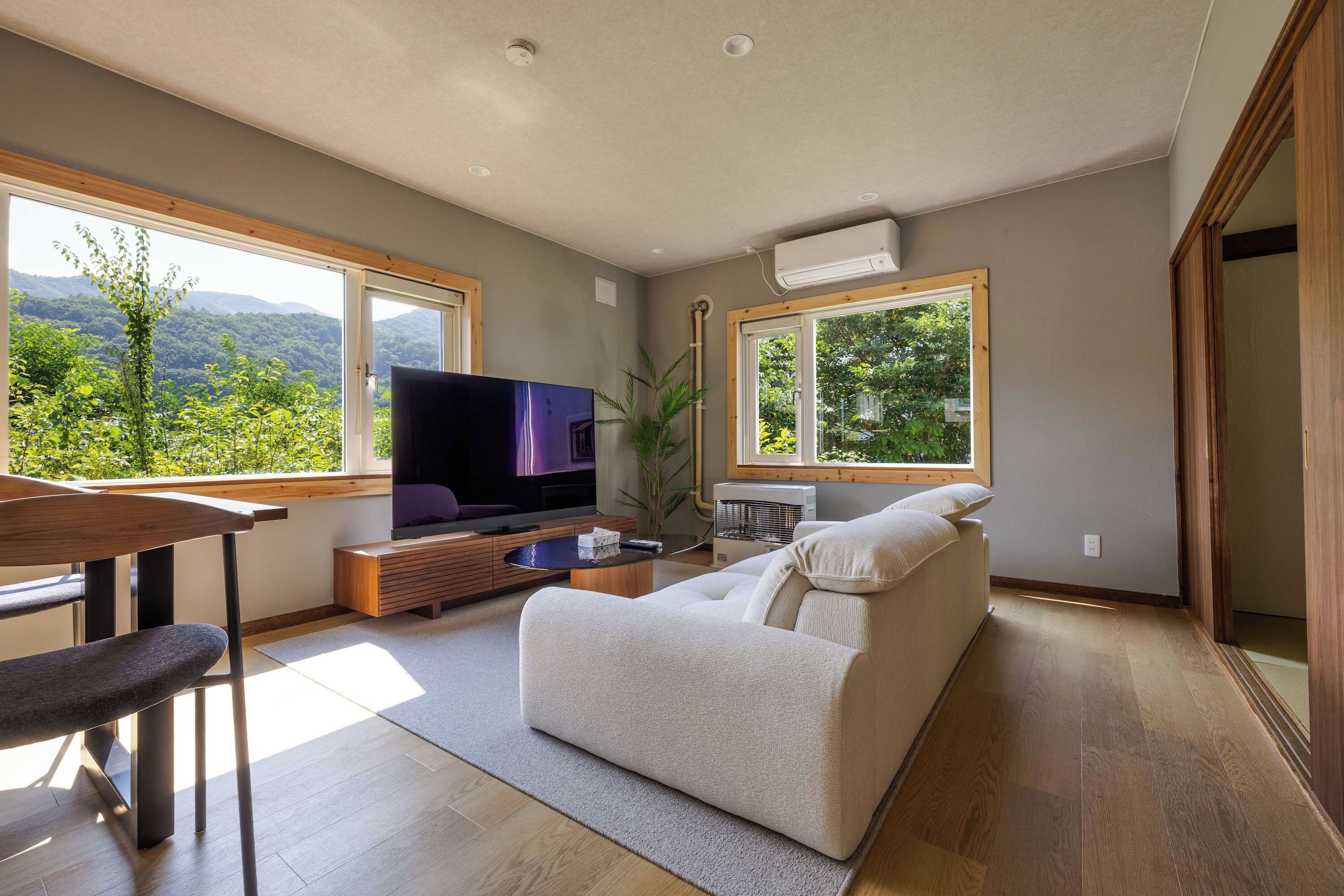 A modern living room with a flat-screen TV, a wooden dining table, a sofa, and large windows offering a view of greenery.