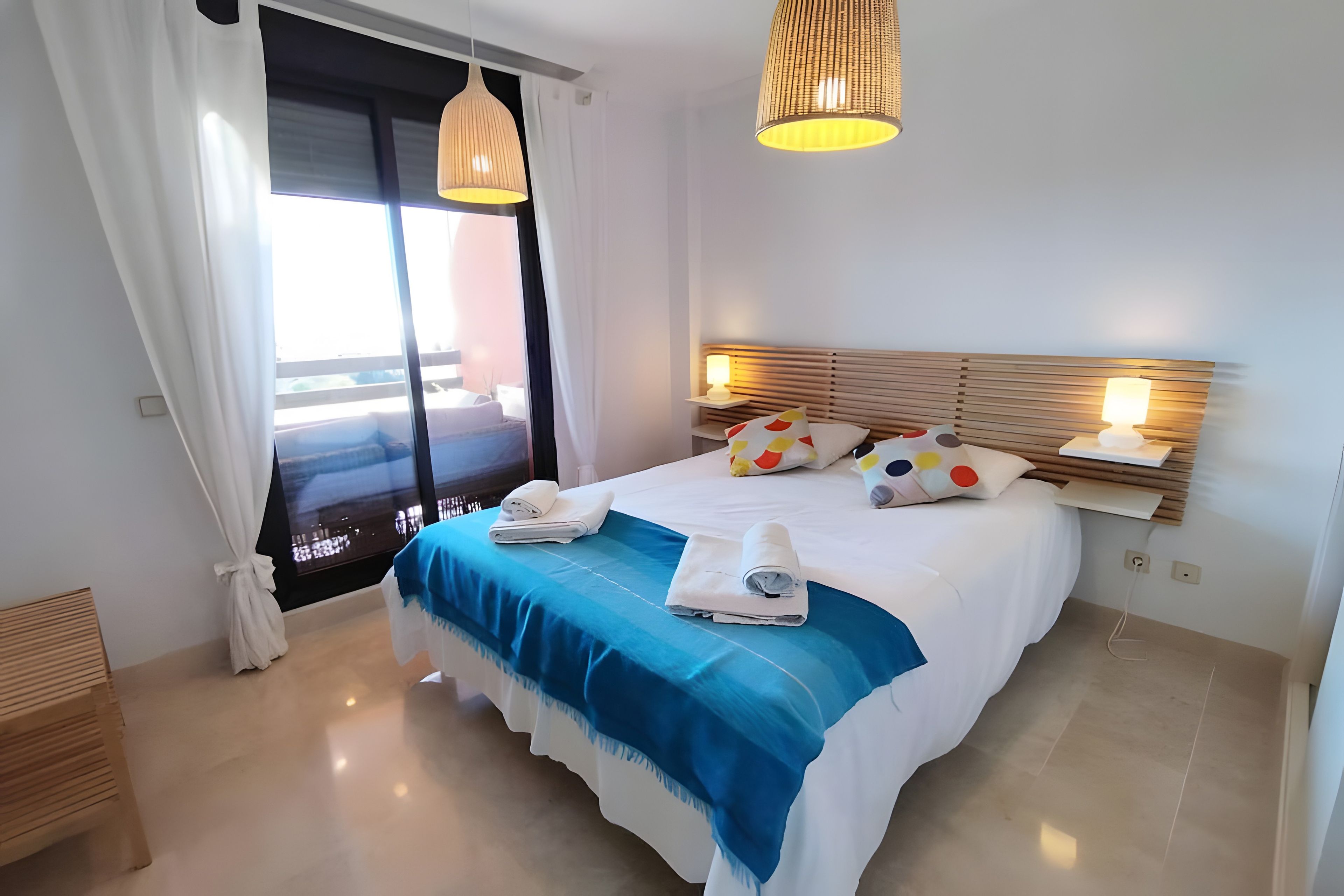 Une chambre d’hôtel avec un grand lit, deux lampes de chevet, une vue sur la mer par la fenêtre et une tête de lit en bois.