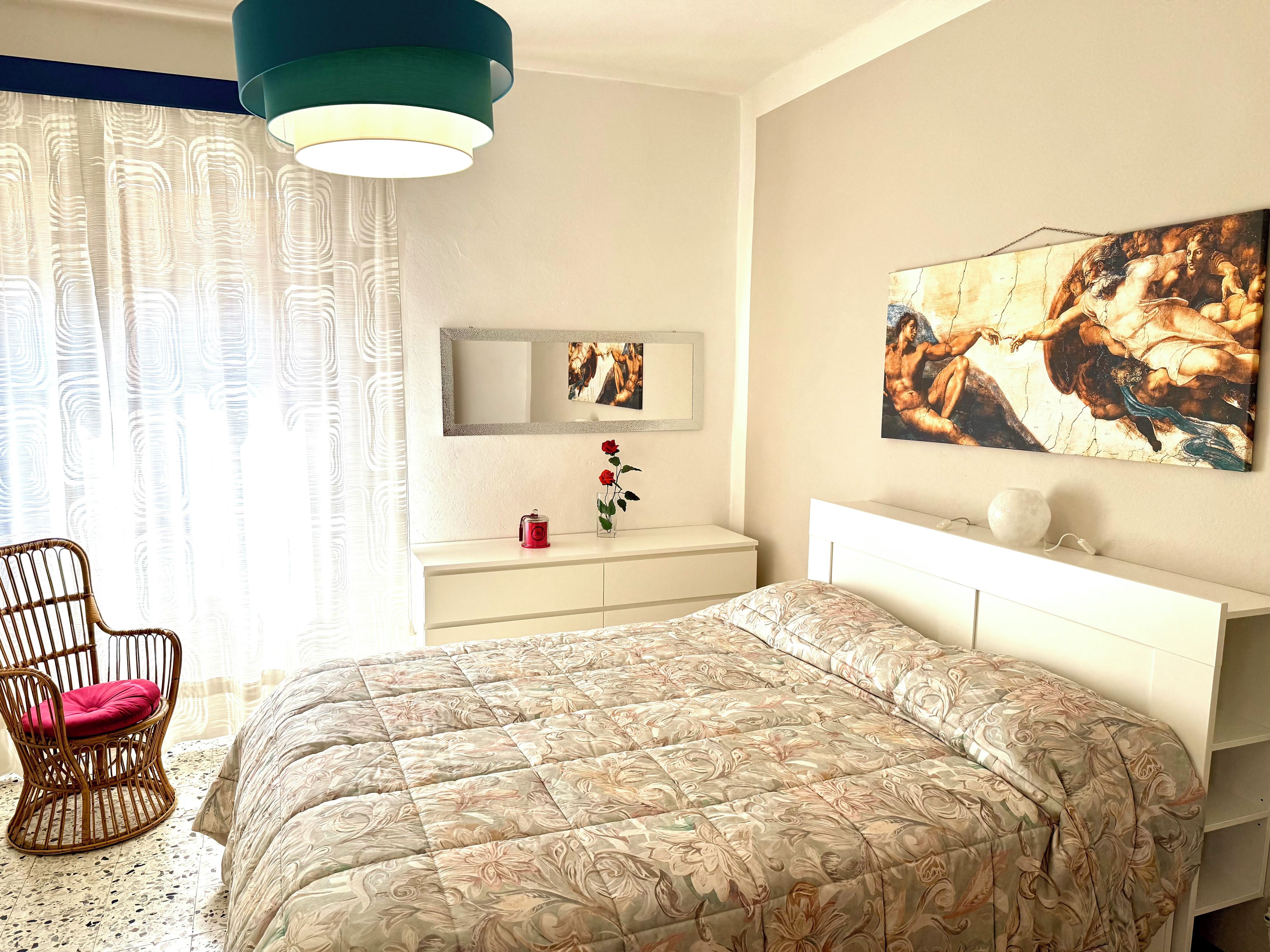 A bedroom with a bed, a chair, a vanity with a mirror, and artwork on the wall.