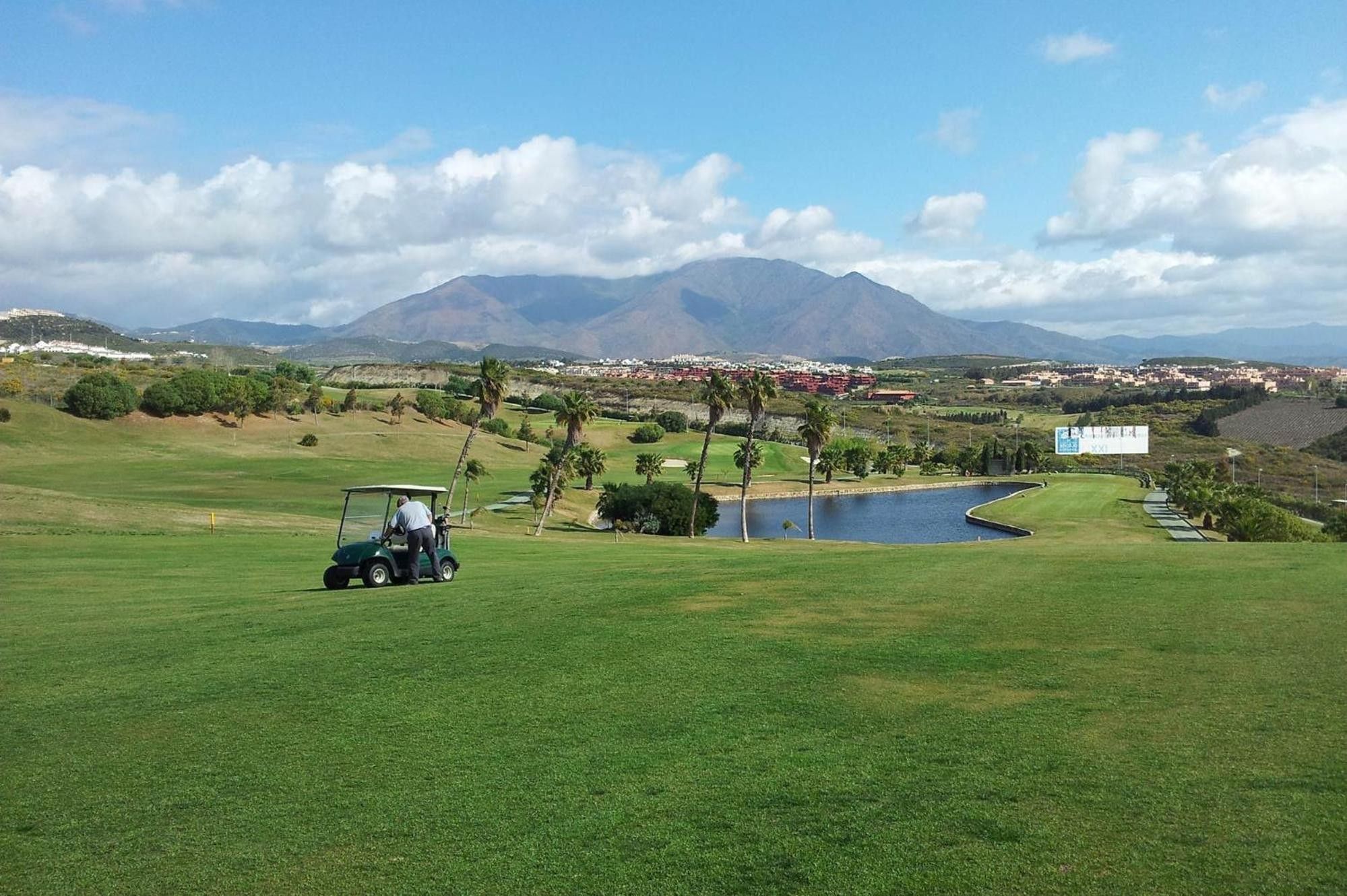 A golf course with a pond, palm trees, and mountains in the background.