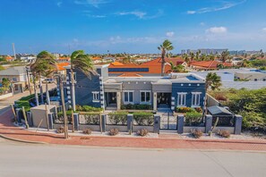 A two-story residential building with a flat roof, surrounded by a landscaped garden with cacti and shrubs. - Spacious 5BR Aruba Retreat with 2 Private Pools, Guest Apartment, 2 BBQ's & Outdoor Lounge (Noord)