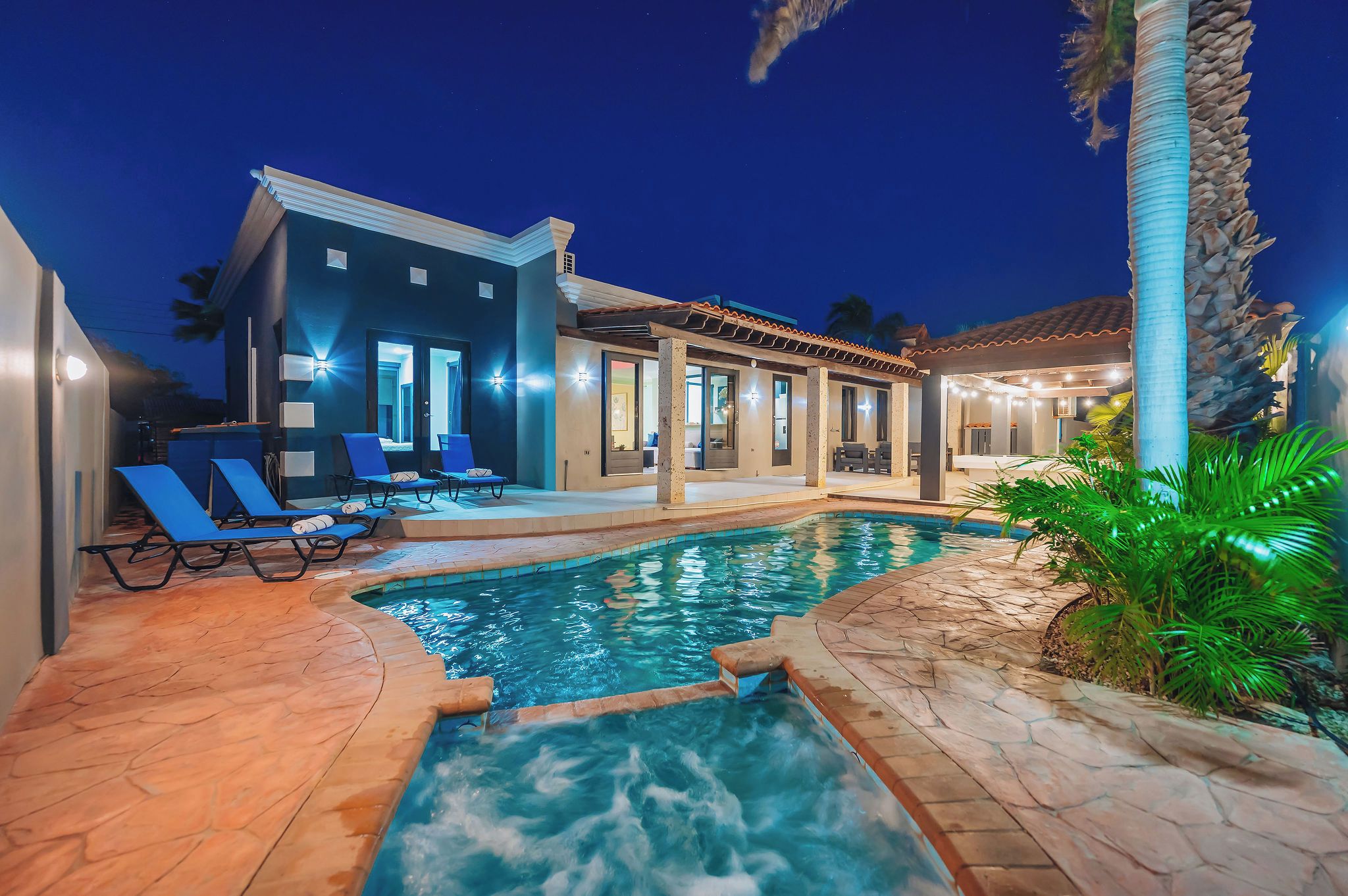 Une piscine avec des chaises longues, un patio couvert et une maison dotée de nombreuses portes et fenêtres.
