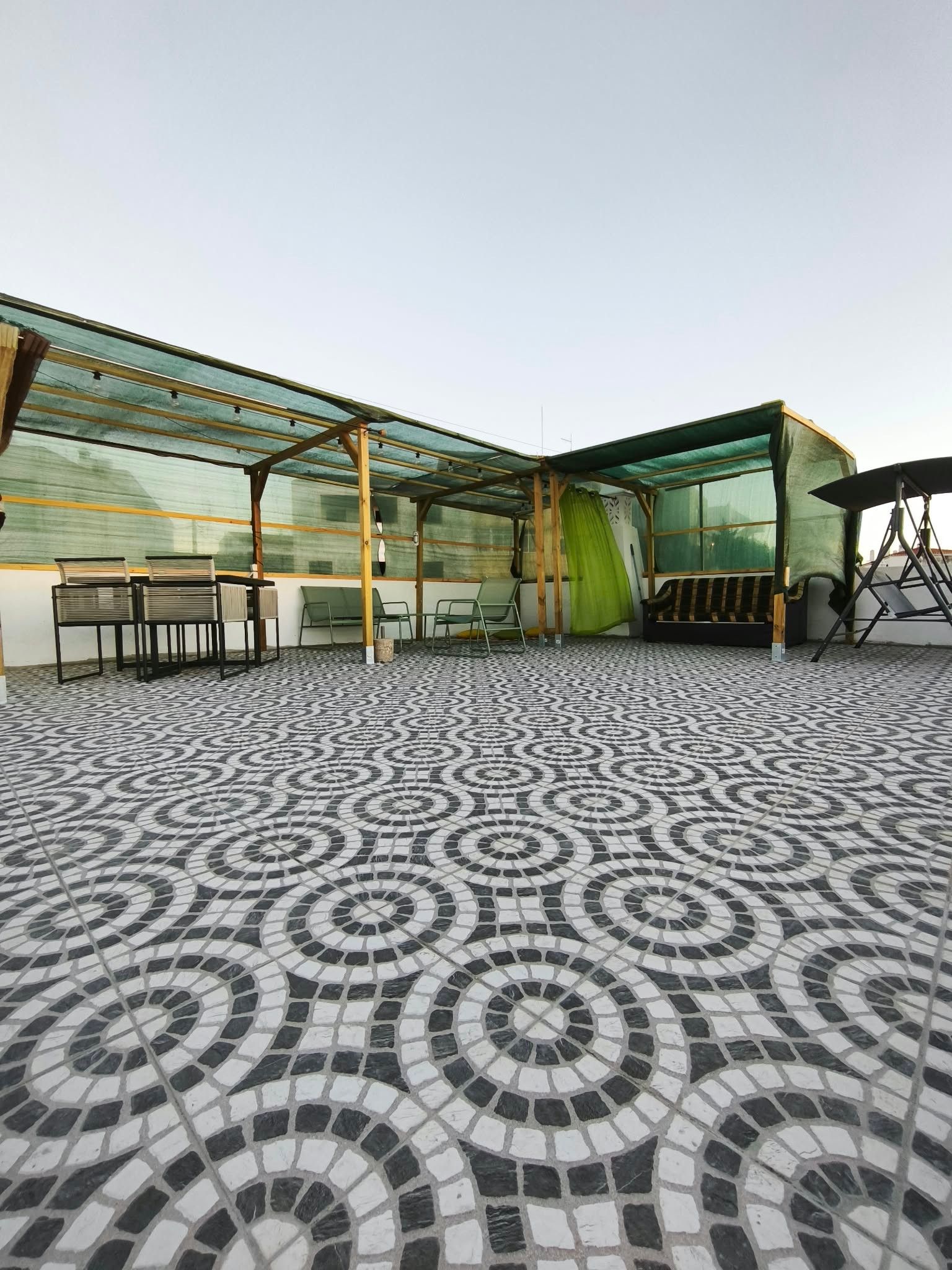 A covered outdoor seating area with patterned floor tiles, a table with chairs, and a green curtain.