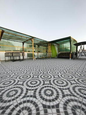A covered outdoor seating area with patterned floor tiles, a table with chairs, and a green curtain.