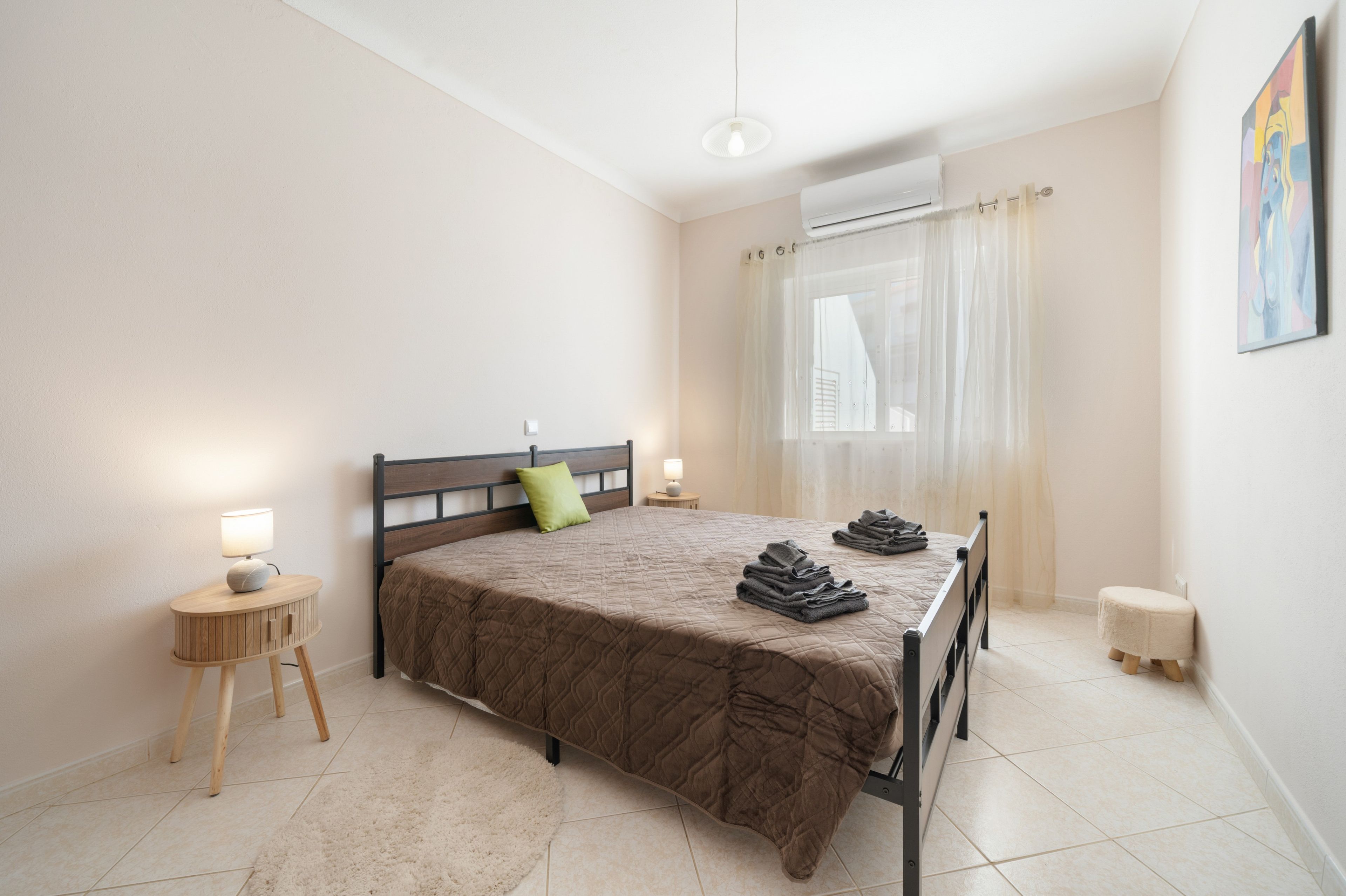 A bedroom with a bed, a nightstand, a lamp, a window with curtains, and a painting on the wall.