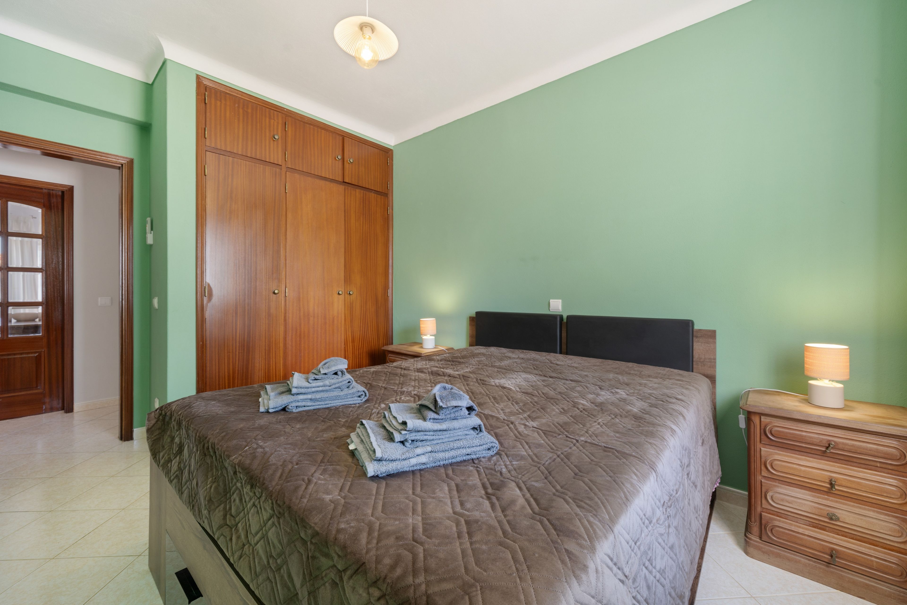 A bedroom with a large bed, wooden wardrobe, and a nightstand with a lamp.