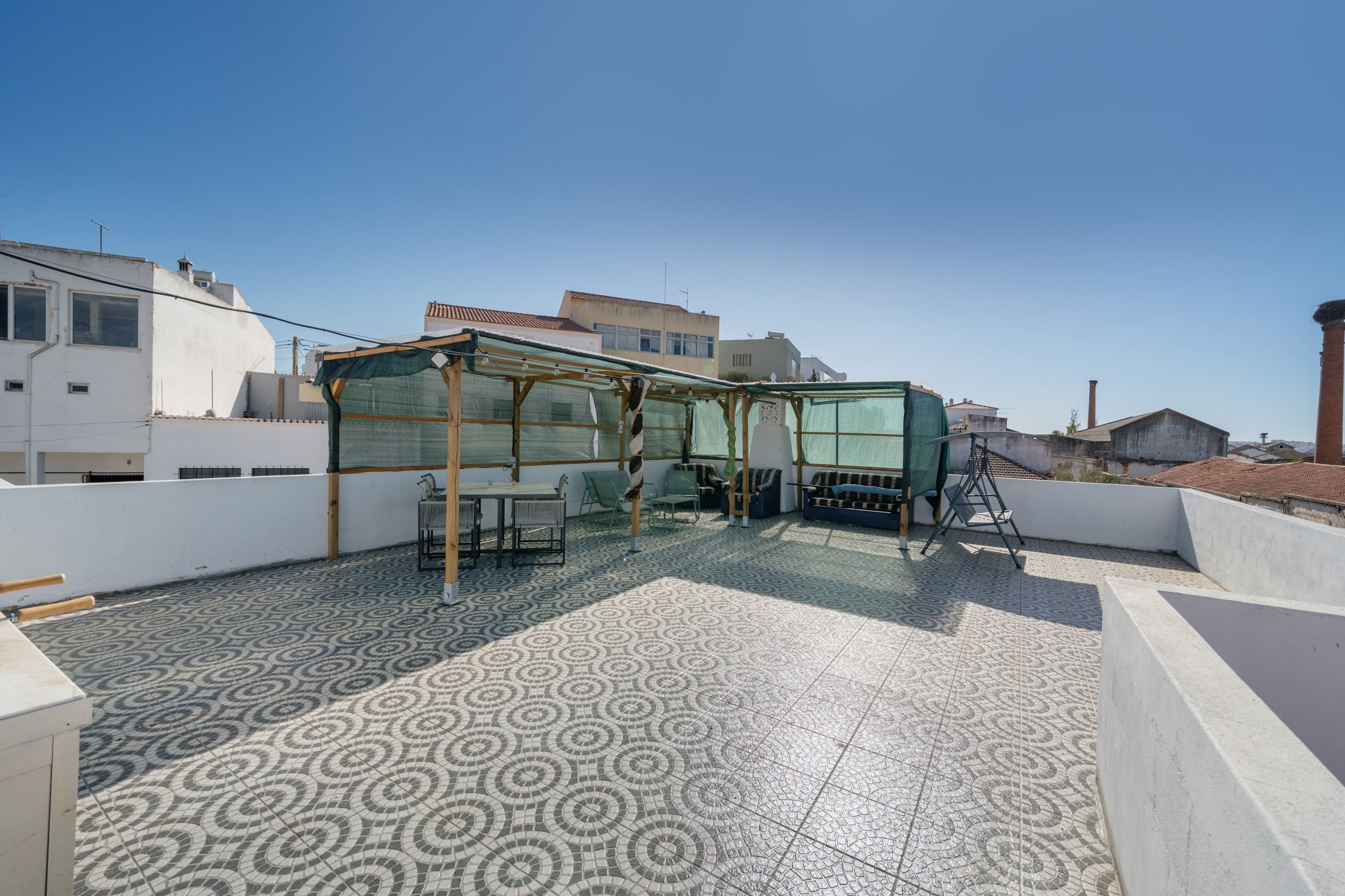 A rooftop terrace with a patterned tile floor, a covered seating area with chairs and a table, and a view of surrounding buildings.