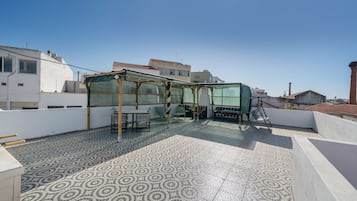 A rooftop terrace with a patterned tile floor, a covered seating area with chairs and a table, and a view of surrounding buildings.