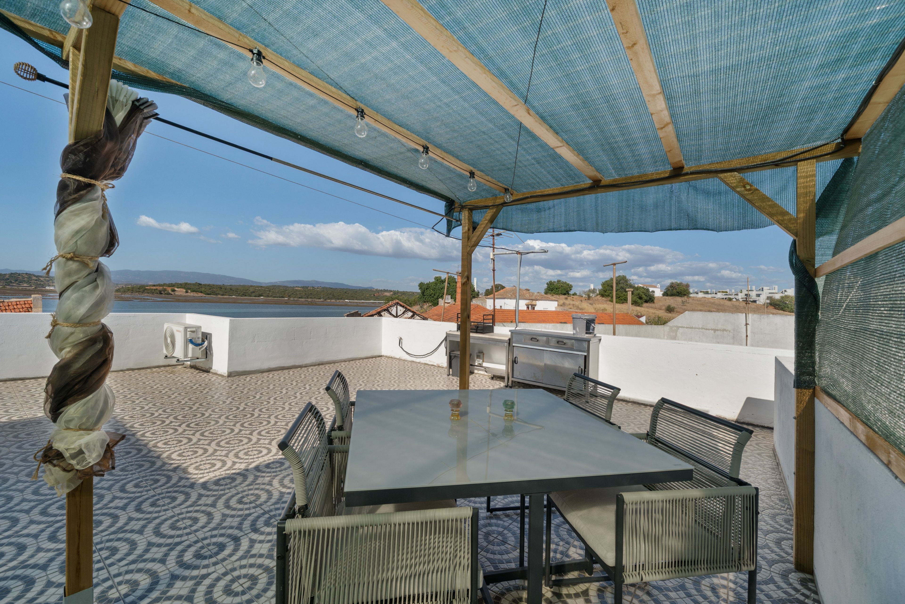 A covered outdoor seating area with a table and chairs, overlooking a body of water and distant hills.