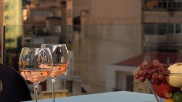 A table with two wine glasses and a fruit bowl on a balcony overlooking a cityscape with buildings and a hill.