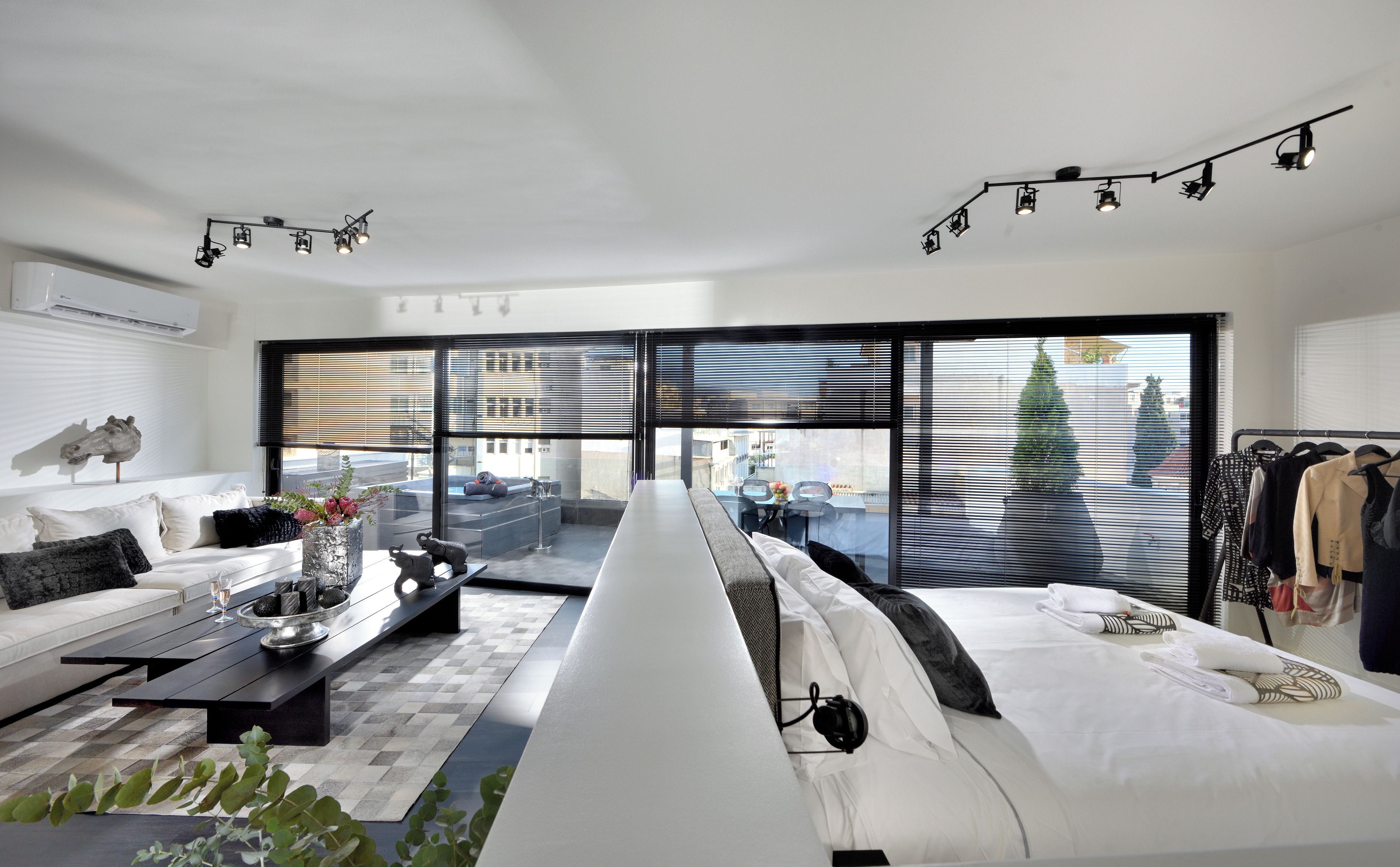 Un salon moderne avec un canapé, une table basse et un coin repas. Il y a une grande fenêtre avec des stores, un luminaire au plafond et une vue sur la ville.
