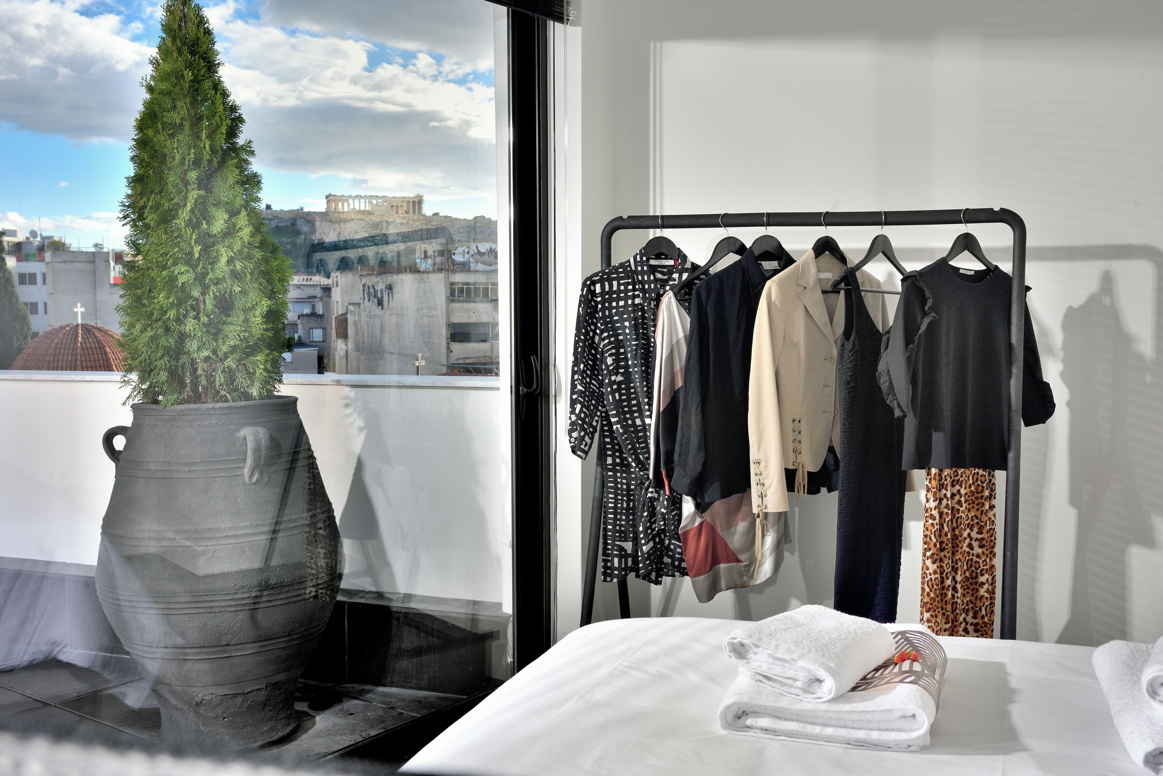 Ein Zimmer mit einem Bett, einem Kleiderständer mit verschiedenen Kleidungsstücken, einer Topfpflanze und Blick auf eine Stadtansicht durch ein Fenster.