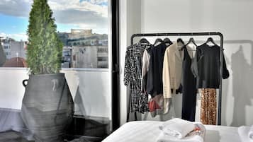 Ein Zimmer mit einem Bett, einem Kleiderständer mit verschiedenen Kleidungsstücken, einer Topfpflanze und Blick auf eine Stadtansicht durch ein Fenster.