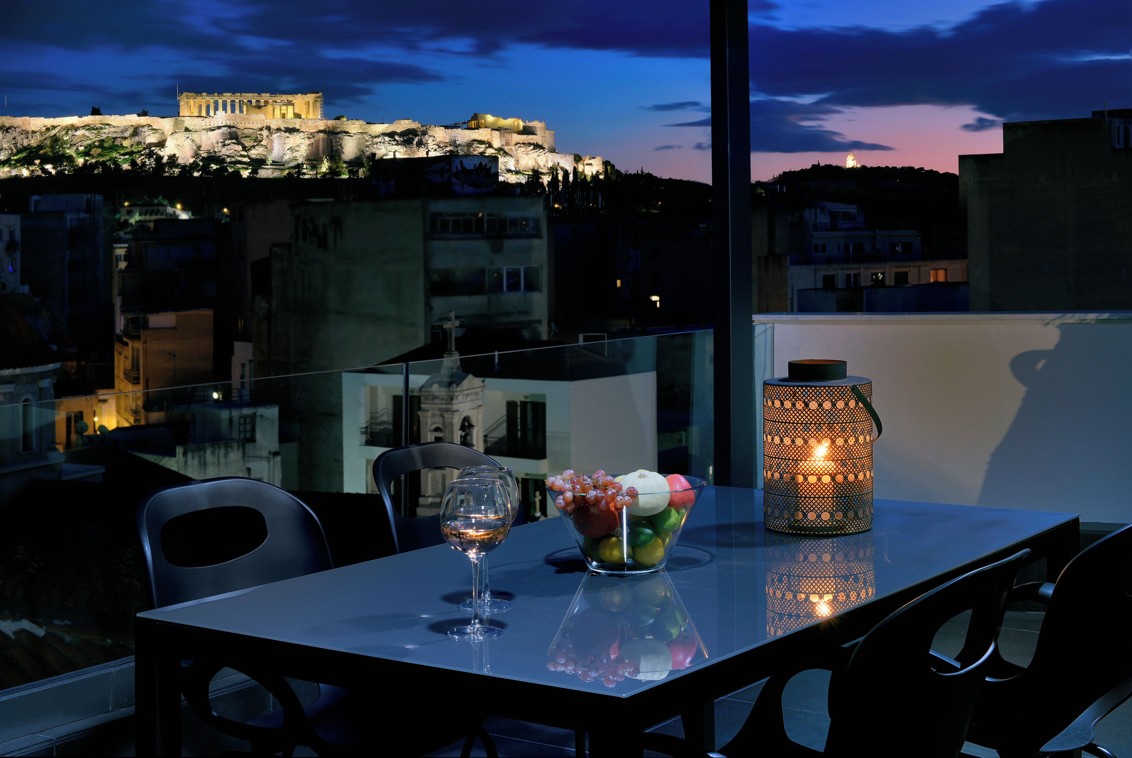 Une table avec un verre à vin, un panier de fruits et une lanterne, sur un balcon donnant sur un paysage urbain avec des bâtiments historiques.