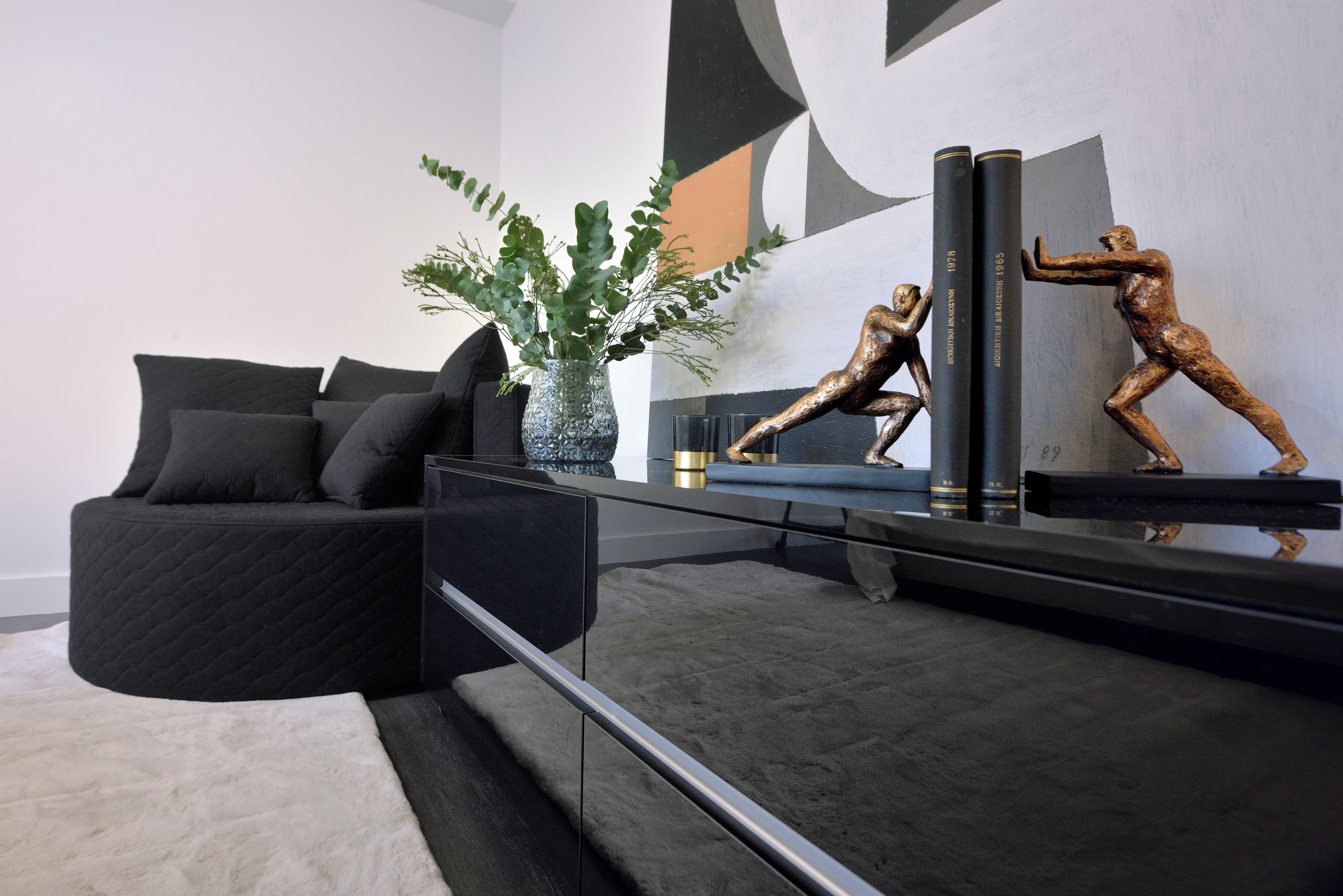 Un salon moderne avec un canapé noir, une table basse en verre et des objets décoratifs, notamment un vase avec des feuilles vertes et deux sculptures en bronze.
