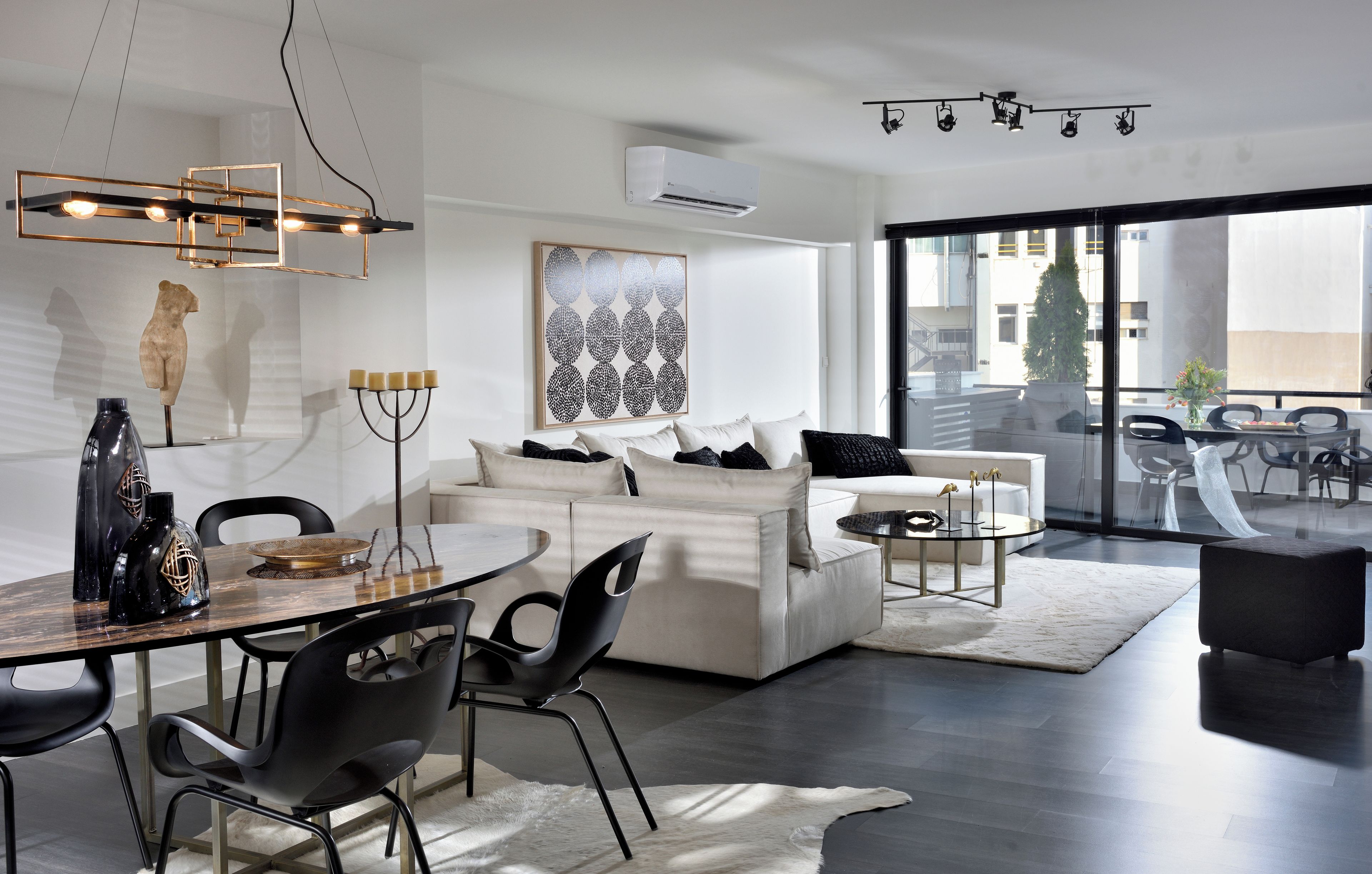 A modern living room with a white sofa, black chairs, a dining table, and a large window offering a view of the city.