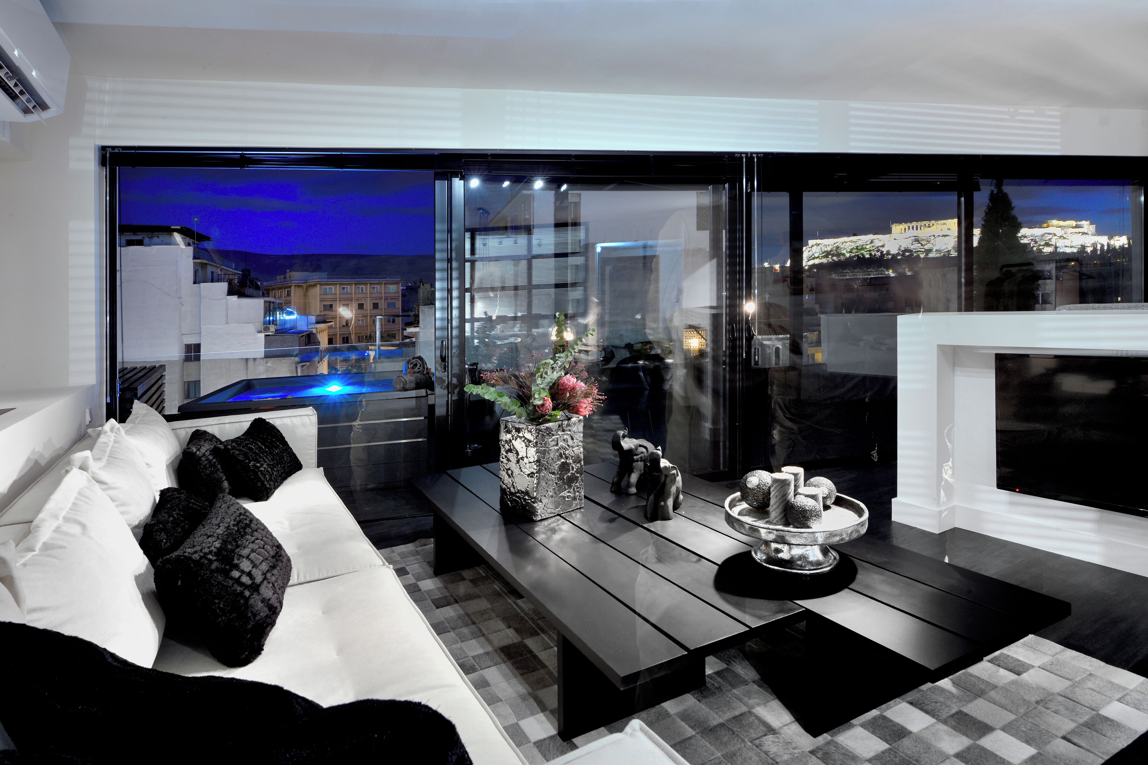 Ein modernes Wohnzimmer mit einer Couch, einem Couchtisch und einem Kamin. Ein großes Fenster bietet Blick auf eine Skyline und einen Pool.