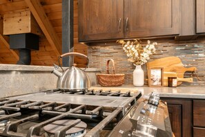 A kitchen with a gas stove, a kettle, a basket, a vase with flowers, and a wooden cutting board.
