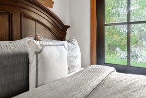 A bed with a white quilt and pillows, a wooden headboard, and a window with a view of greenery.