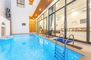 A modern indoor swimming pool with clear glass walls, a wooden ceiling, and a seating area with a sofa and chairs. - Villa C | 6BR Private Pool | Central Pattaya (Pattaya City)