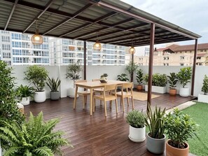 Een terras buiten met een houten tafel en stoelen, omringd door potplanten en een overdekt gedeelte met hanglampen.