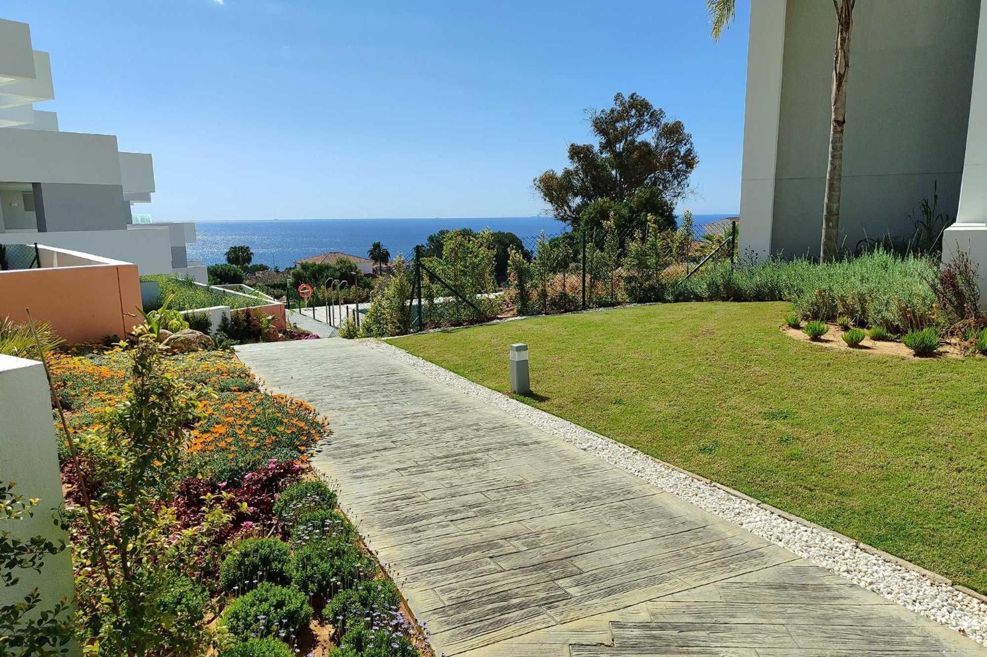 Un sentier qui mène à un espace verdoyant offrant une vue sur la mer, entouré de végétation et d’une architecture moderne.