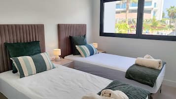 A hotel room with two beds, a window with a view of buildings and greenery, and a nightstand with a lamp.