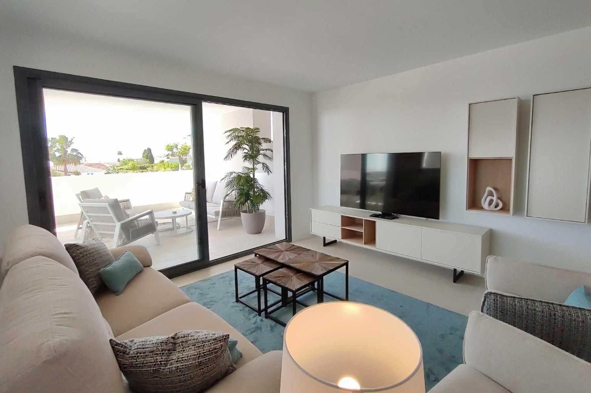 A modern living room with a sofa, a coffee table, a TV, and a sliding glass door leading to an outdoor seating area.