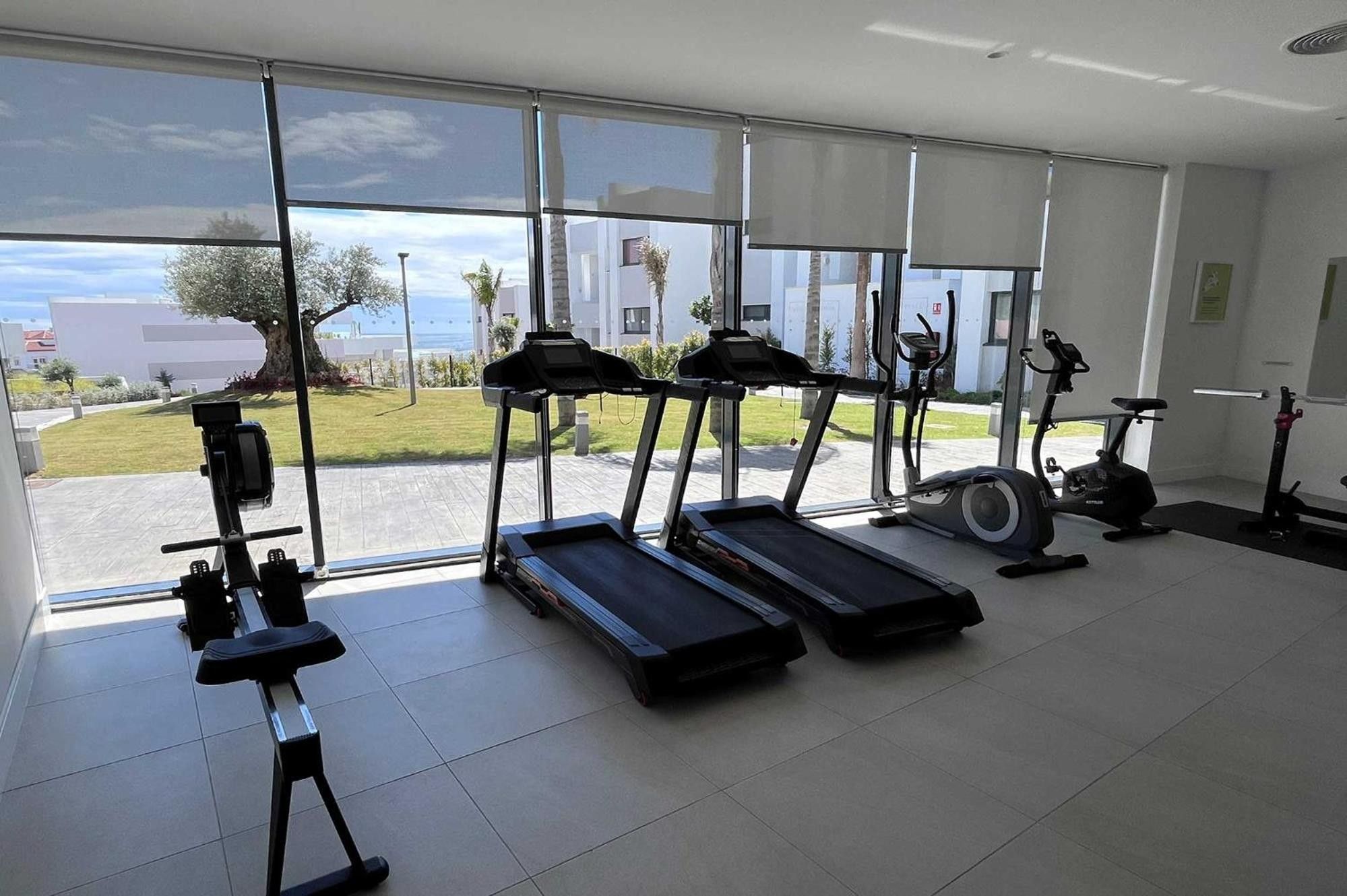Une salle de sport équipée de tapis de course, de vélos elliptiques et d’un rameur, avec de grandes fenêtres donnant sur un bâtiment et sur la verdure.