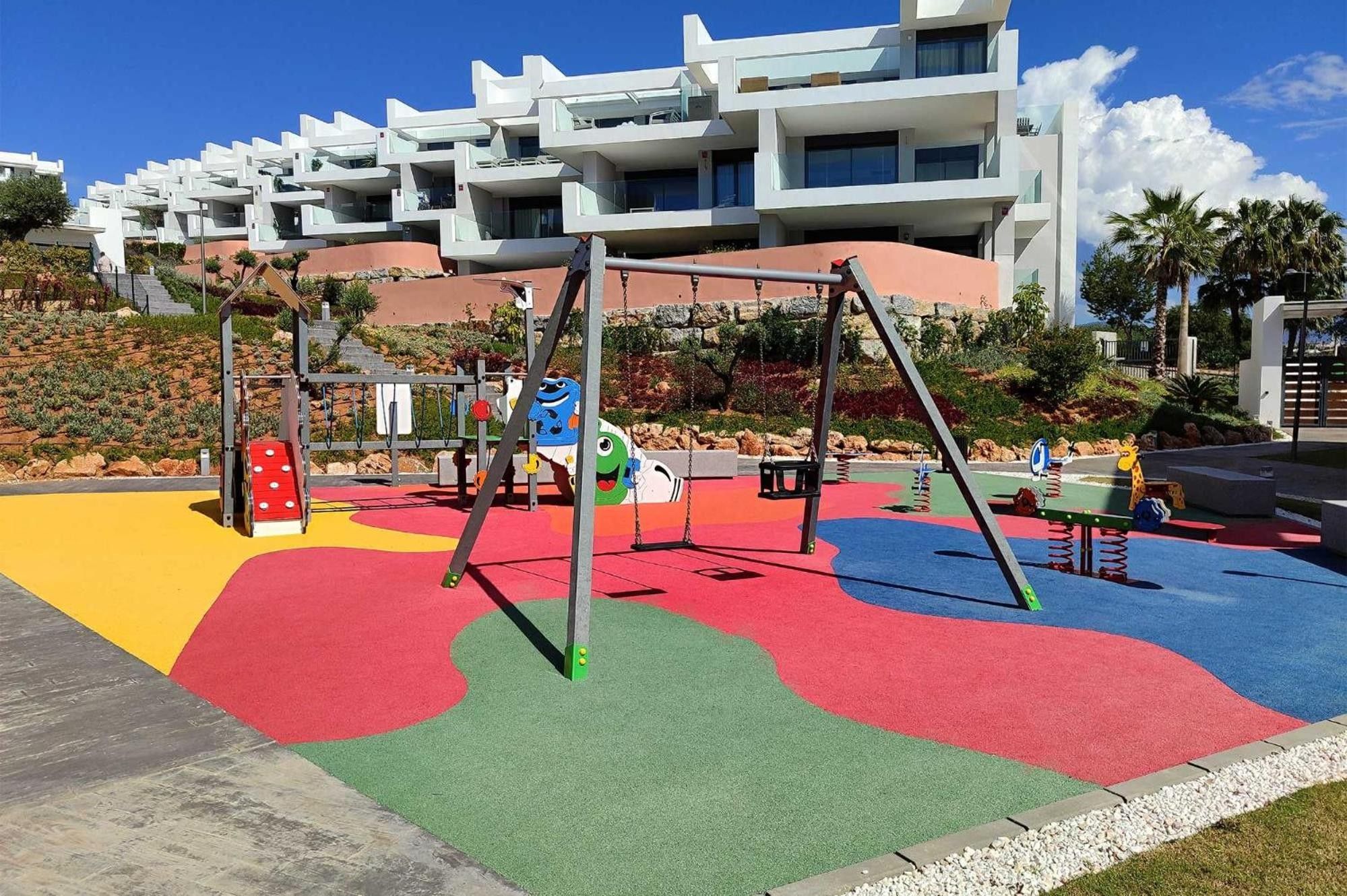 Un terrain de jeux avec des aires de jeux colorées et des balançoires, le tout situé devant un décor composé d’immeubles modernes.