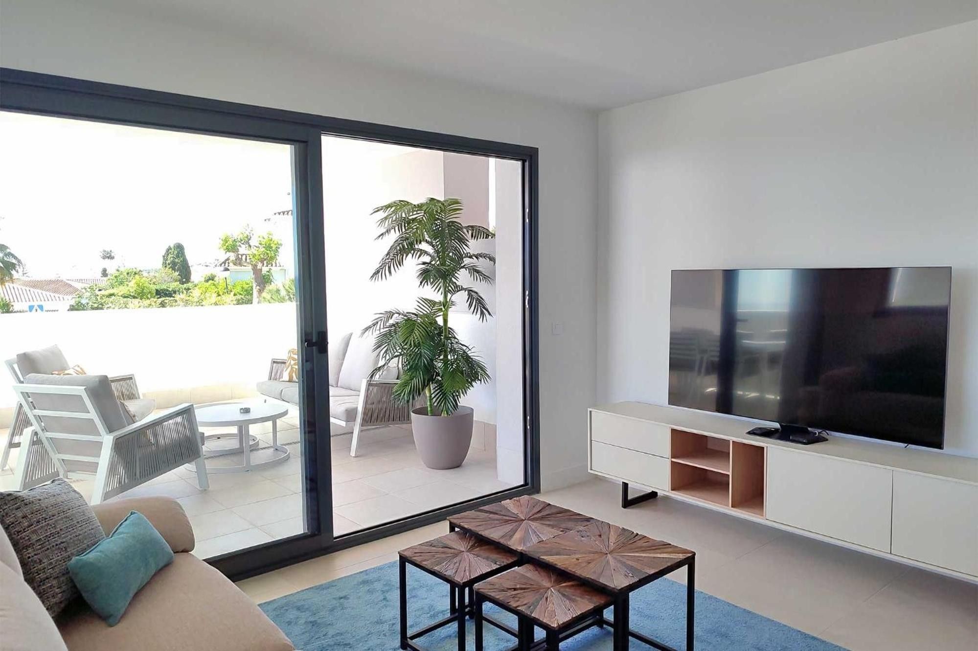 Un salon moderne comprenant un canapé, une table basse, un téléviseur et une porte-fenêtre coulissante donnant sur un espace extérieur agrémenté d’une plante et de mobilier.