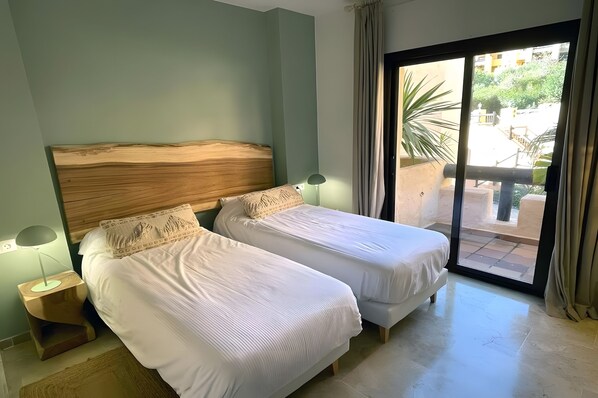Een kamer met twee eenpersoonsbedden, een nachtkastje met een lamp en uitzicht door een glazen deur op een buitenruimte.