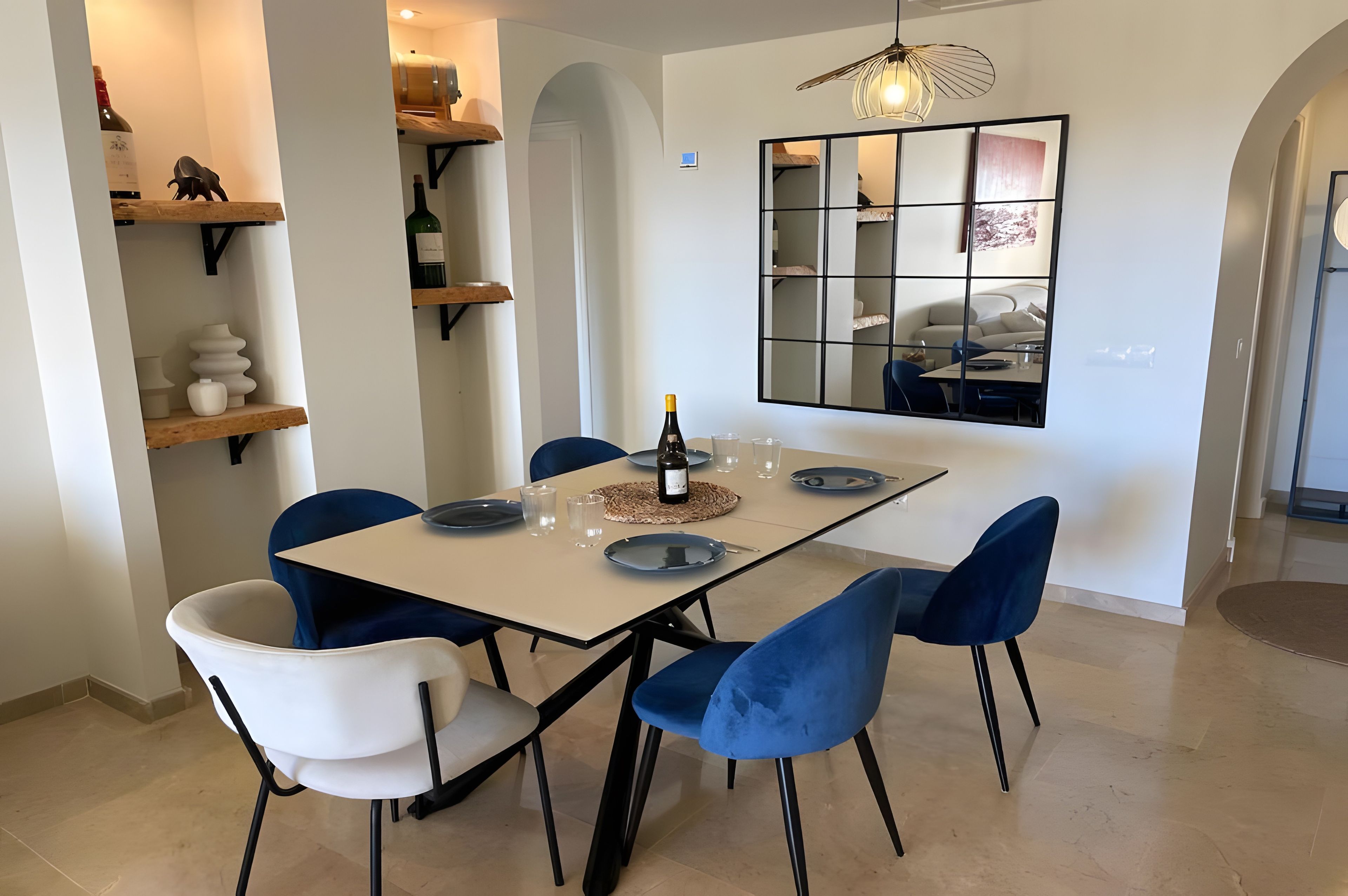 A dining area with a table set for four, blue and white chairs, a wine bottle, and a decorative mirror.