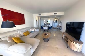 Una sala de estar moderna con un sofá, una mesa de centro y un televisor. Se observa un comedor al fondo.