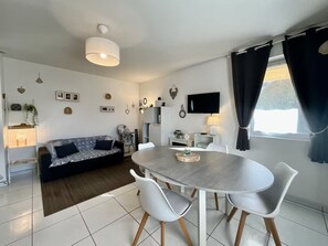 Ein modernes Wohnzimmer mit einer Couch, einem Esstisch, Stühlen, einem Fernseher und einem Fenster mit Vorhängen.