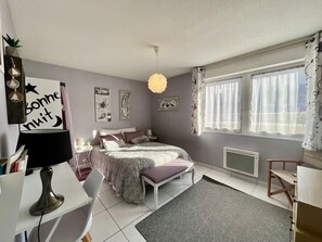 Ein Schlafzimmer mit Bett, Nachttischen, Schreibtisch, Stuhl, einem Fenster mit Vorhängen und Wandbildern.