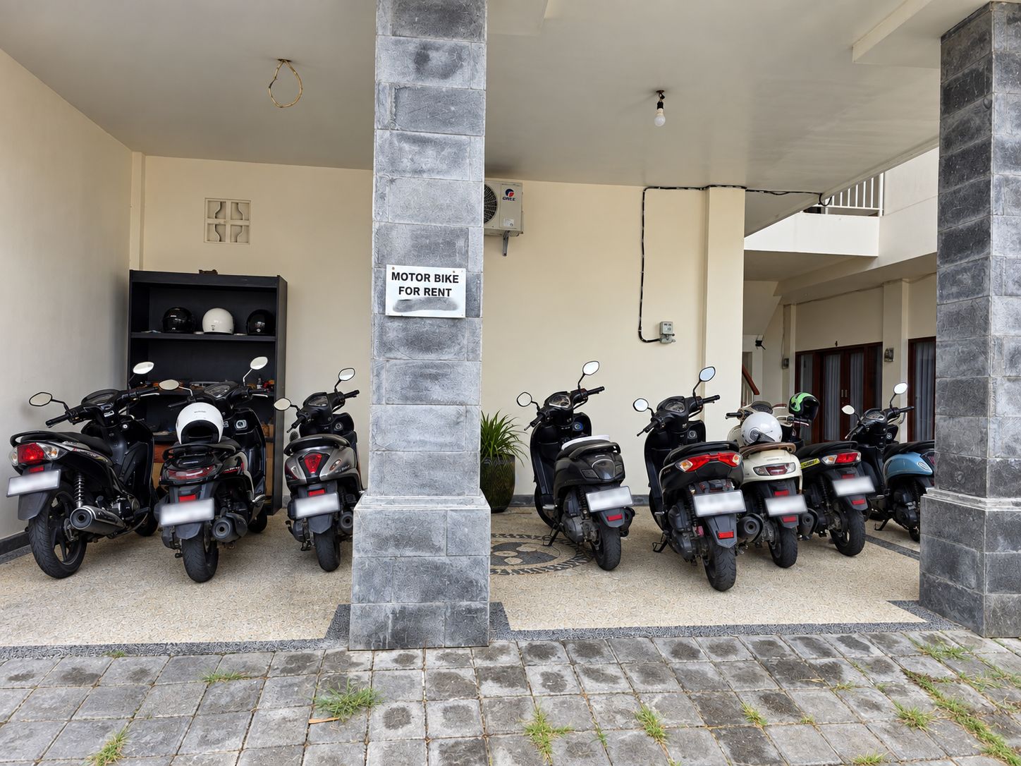 Señal de alquiler de motocicletas, varias scooters estacionadas en un área techada y la entrada a un edificio.