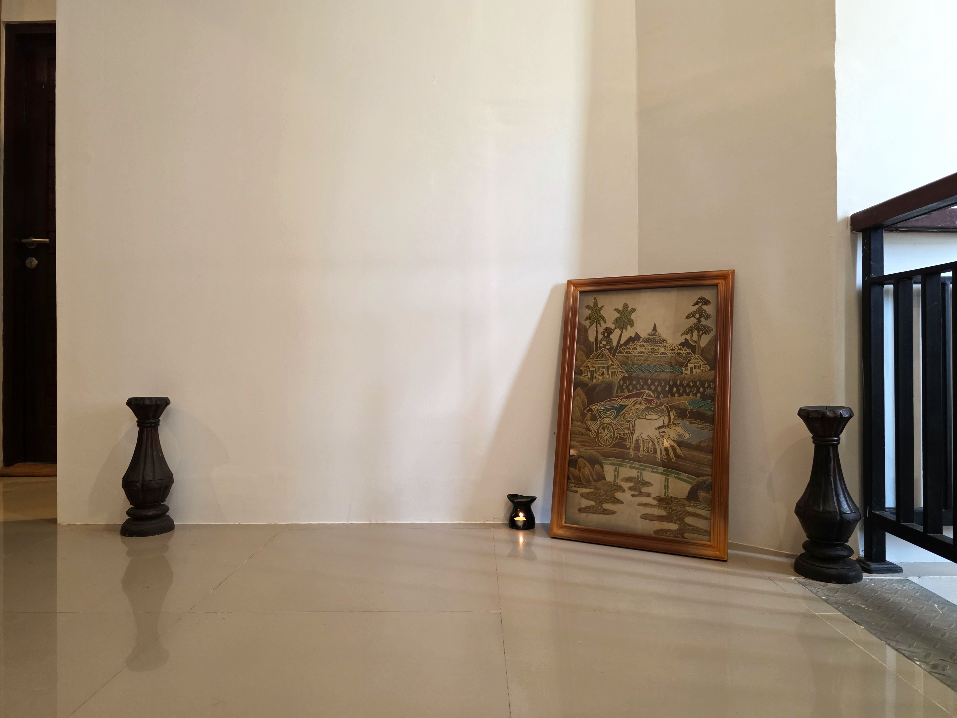 Un pasillo con una obra enmarcada que muestra una escena campestre, una vela en el suelo y dos jarrones a cada lado.