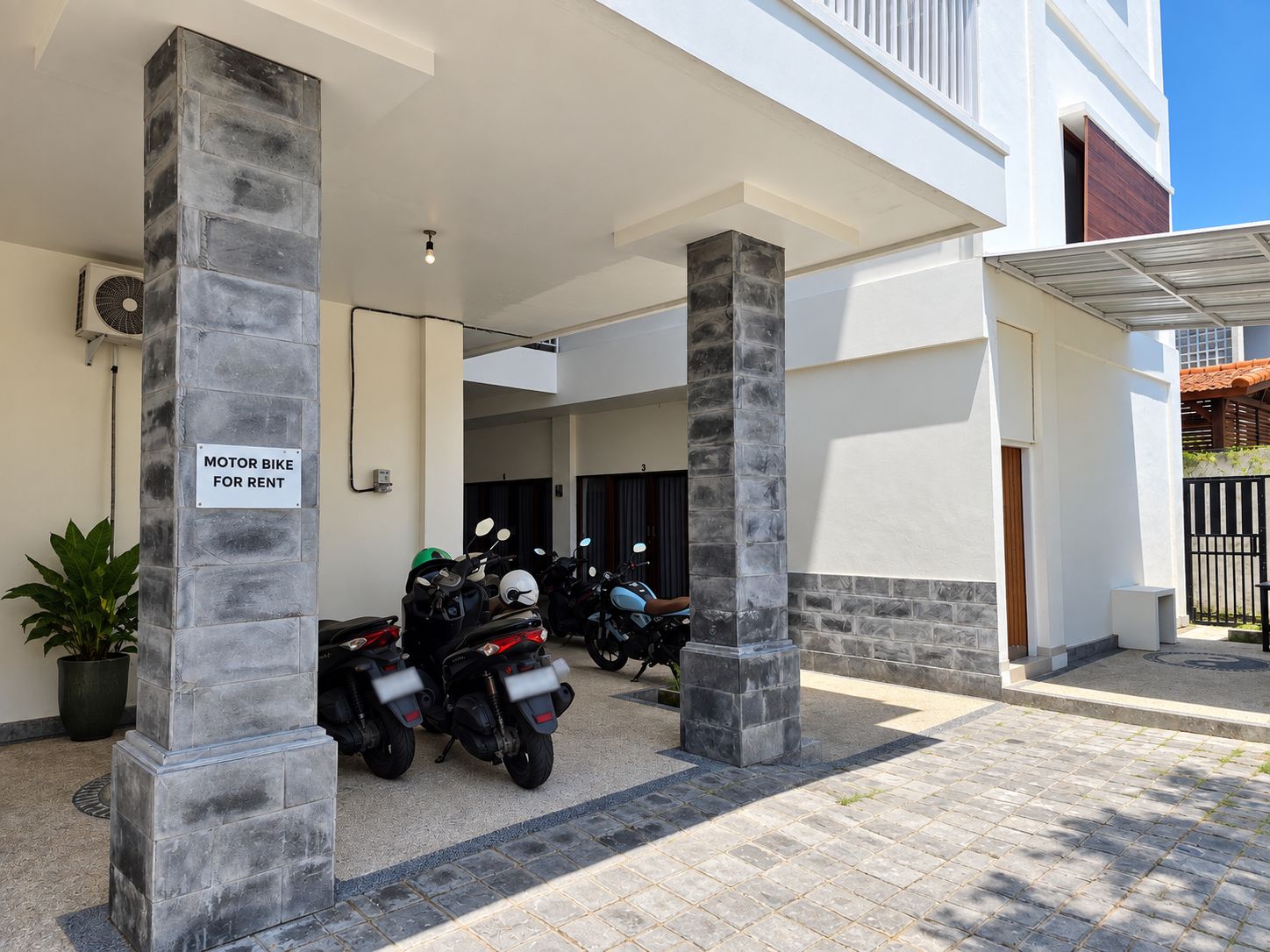 Un área de estacionamiento techada con motos de alquiler, una planta en maceta y un cartel que indica la disponibilidad de motos de alquiler.