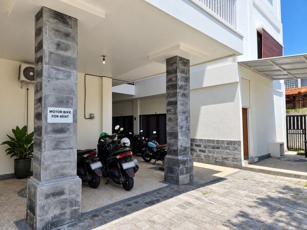A covered parking area with motorbikes for rent, a potted plant, and a sign indicating the availability of motorbikes for rent.