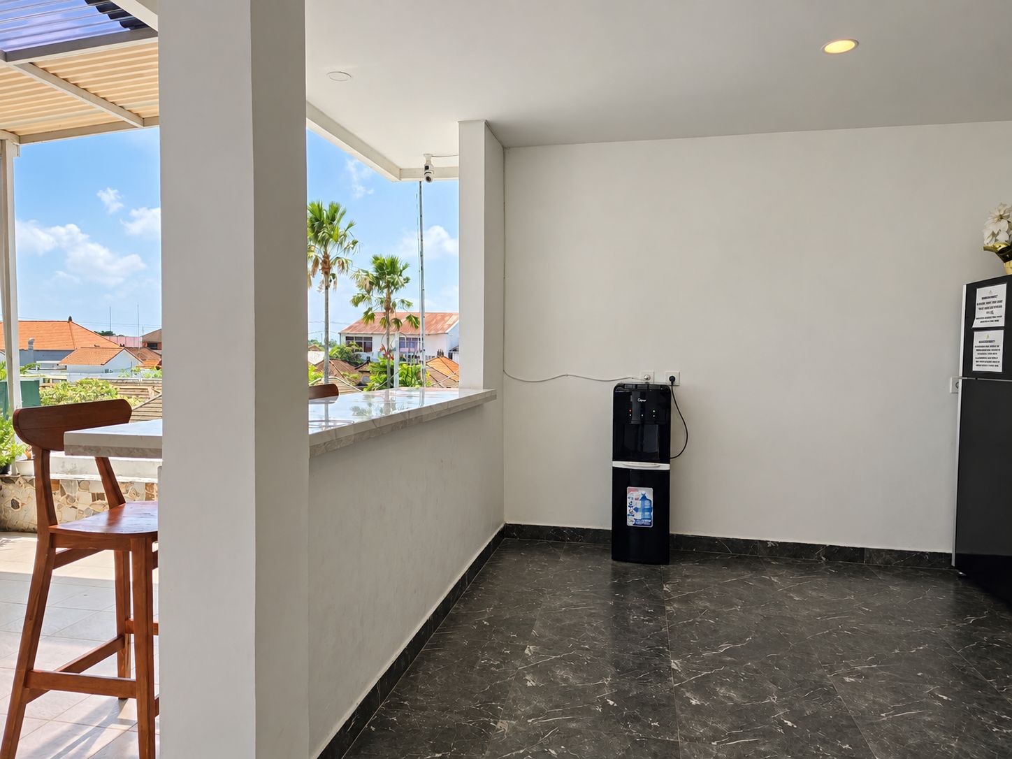 Una habitación con piso de mármol, dispensador de agua, ventana con vista a palmeras y casas, y una silla de madera.