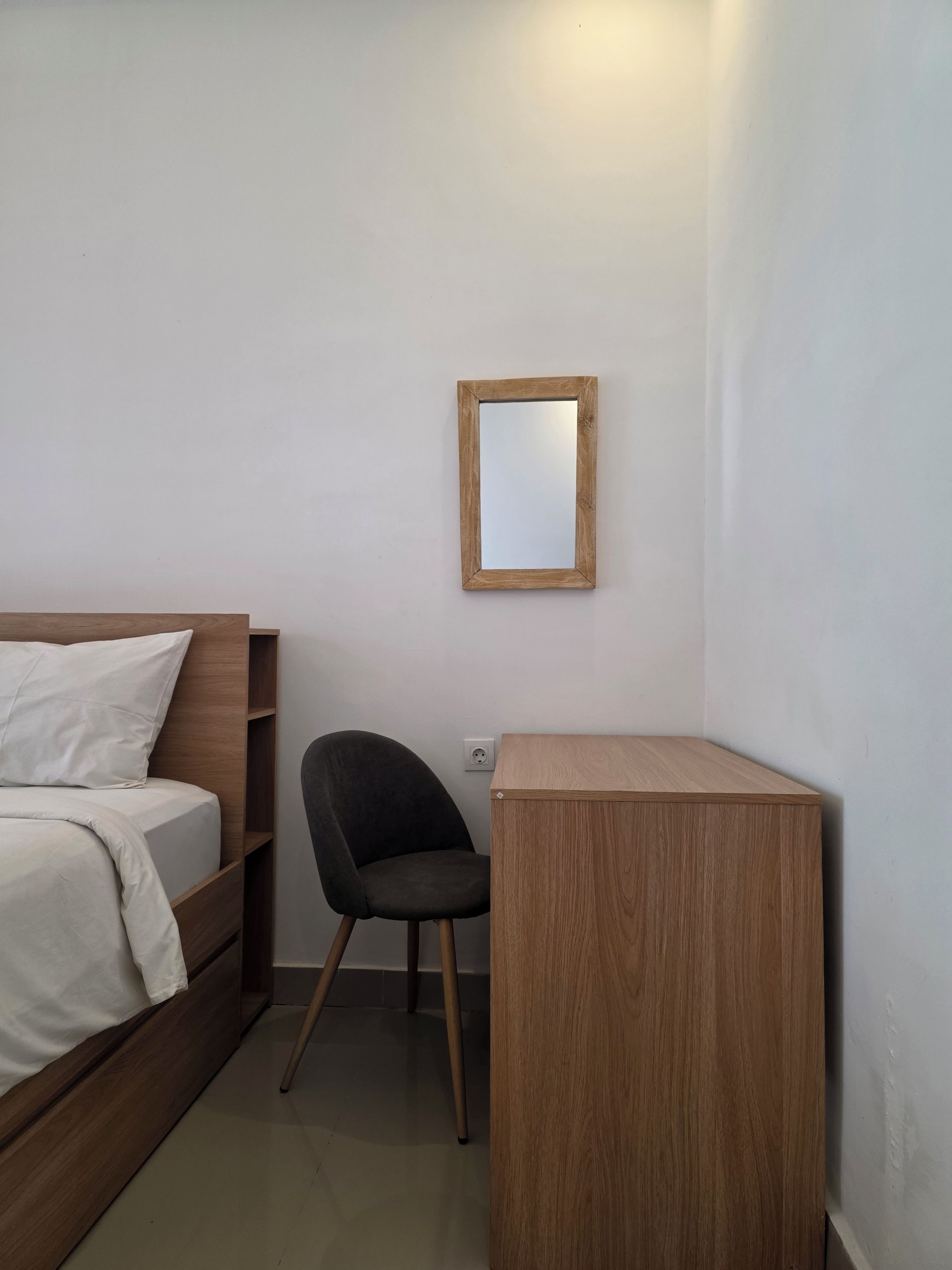 Un dormitorio con una cama de madera, una silla negra, un escritorio de madera pequeño y un espejo en la pared.