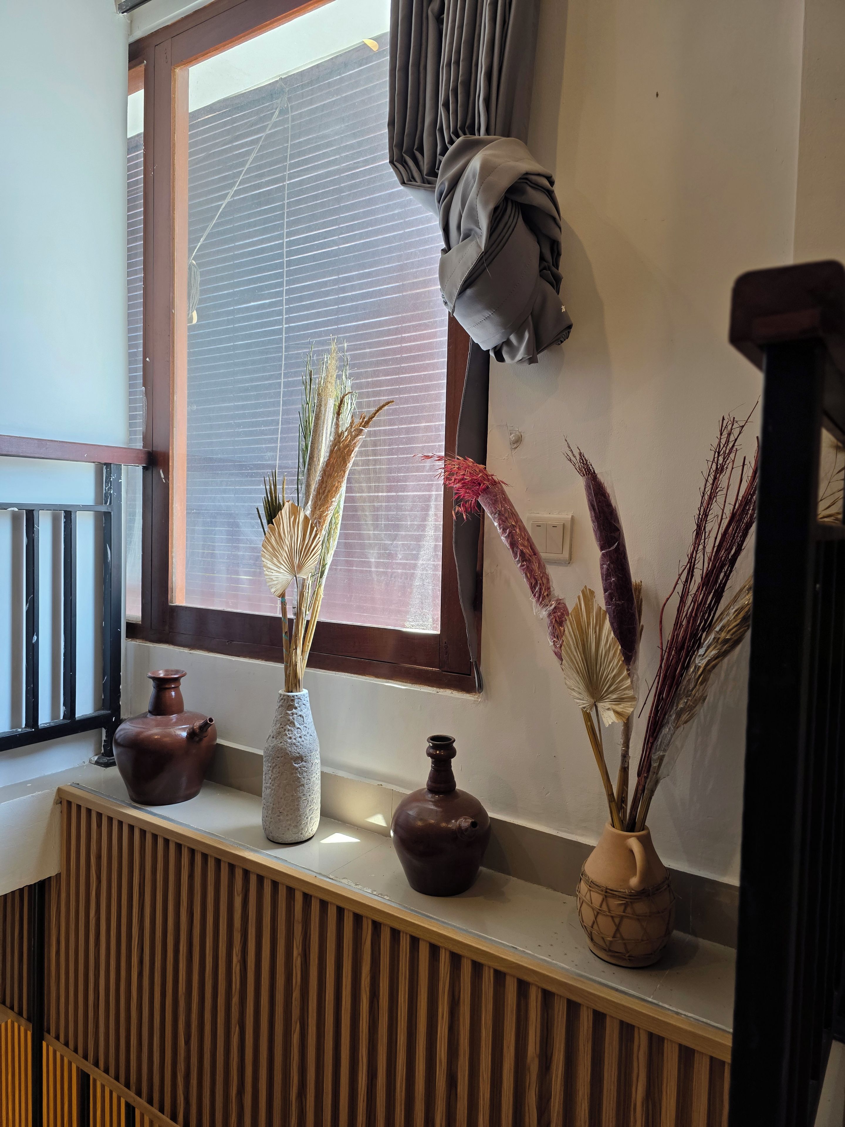 Una ventana con persianas, una barandilla de madera y tres macetas de cerámica con plantas secas en un alféizar.
