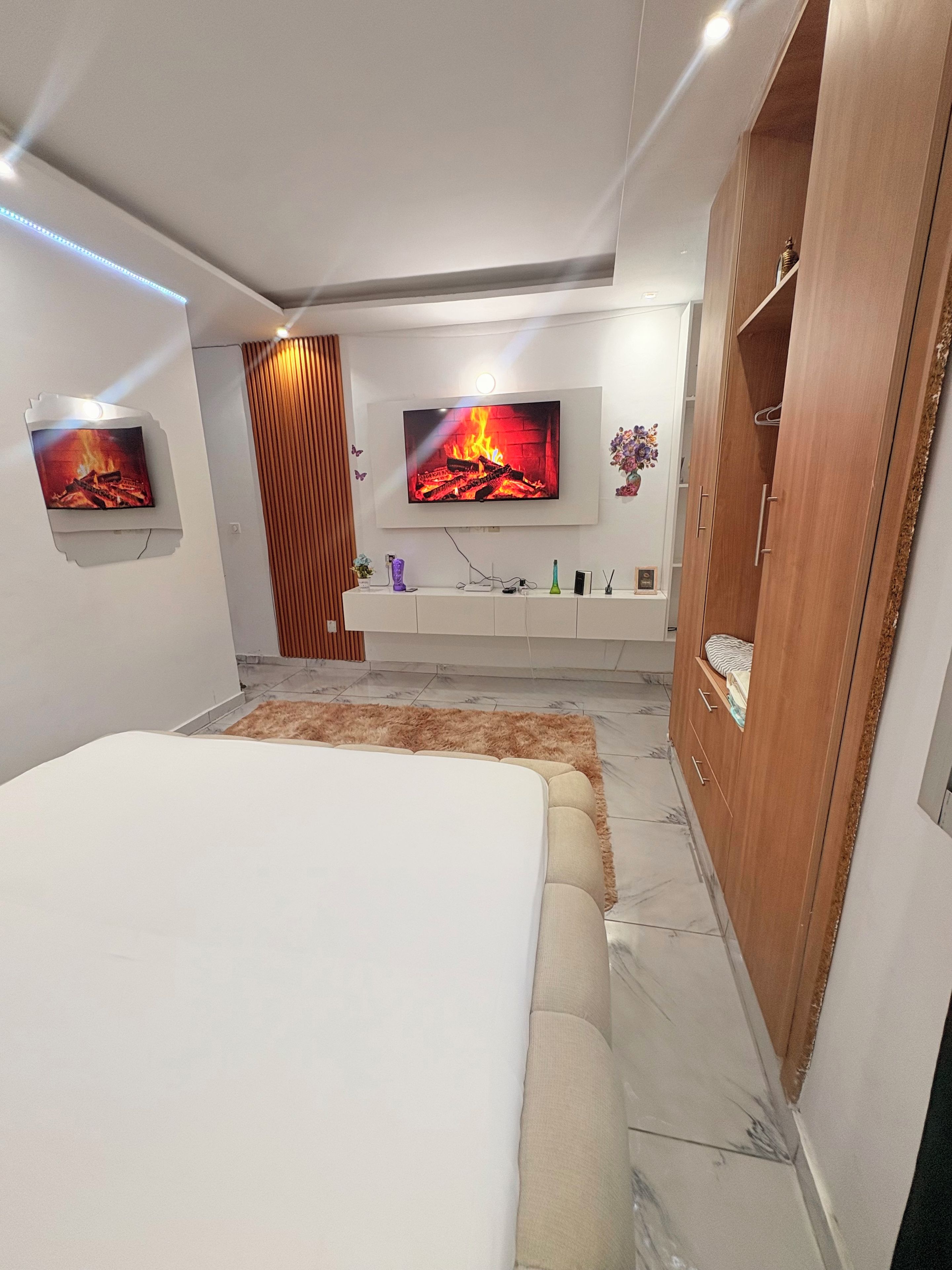 A modern bedroom with a large bed, a TV mounted on the wall, a wardrobe, and a small side table.