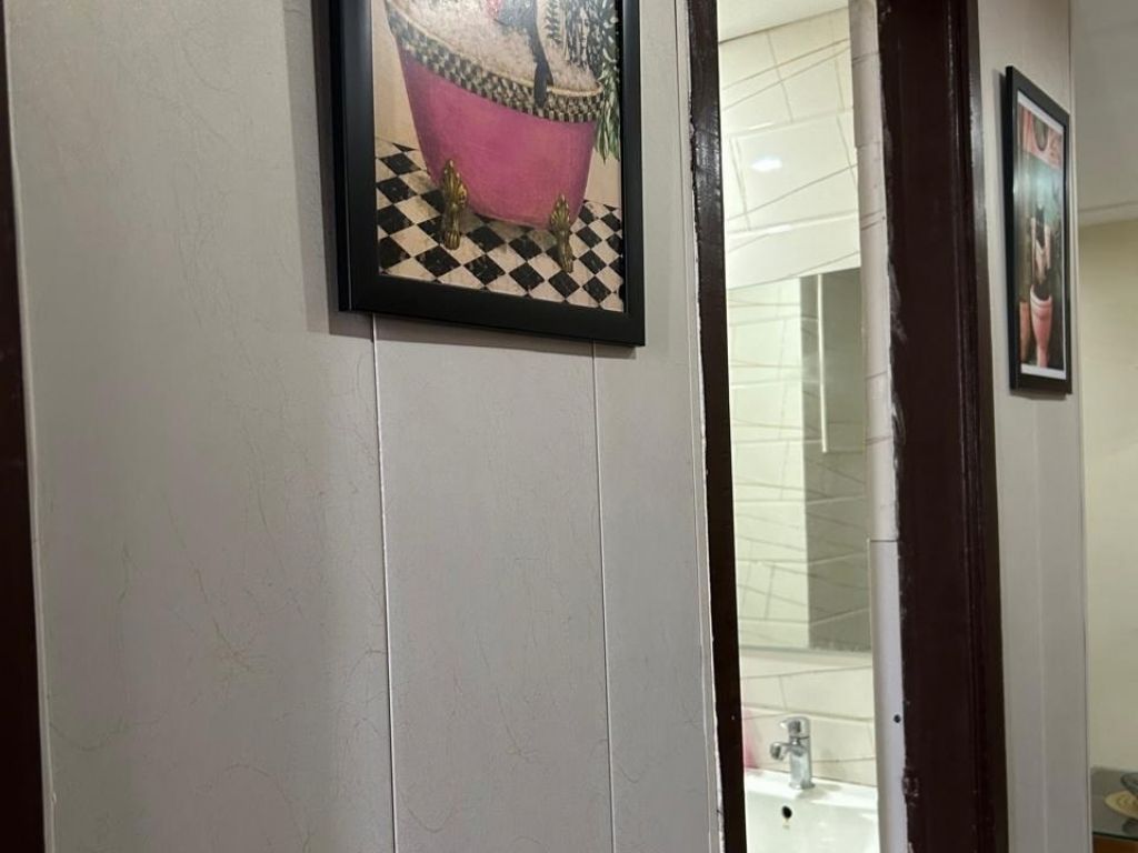A hallway with a framed picture of a pink bathtub on a checkered floor, a white wall, and a door leading to a bathroom.